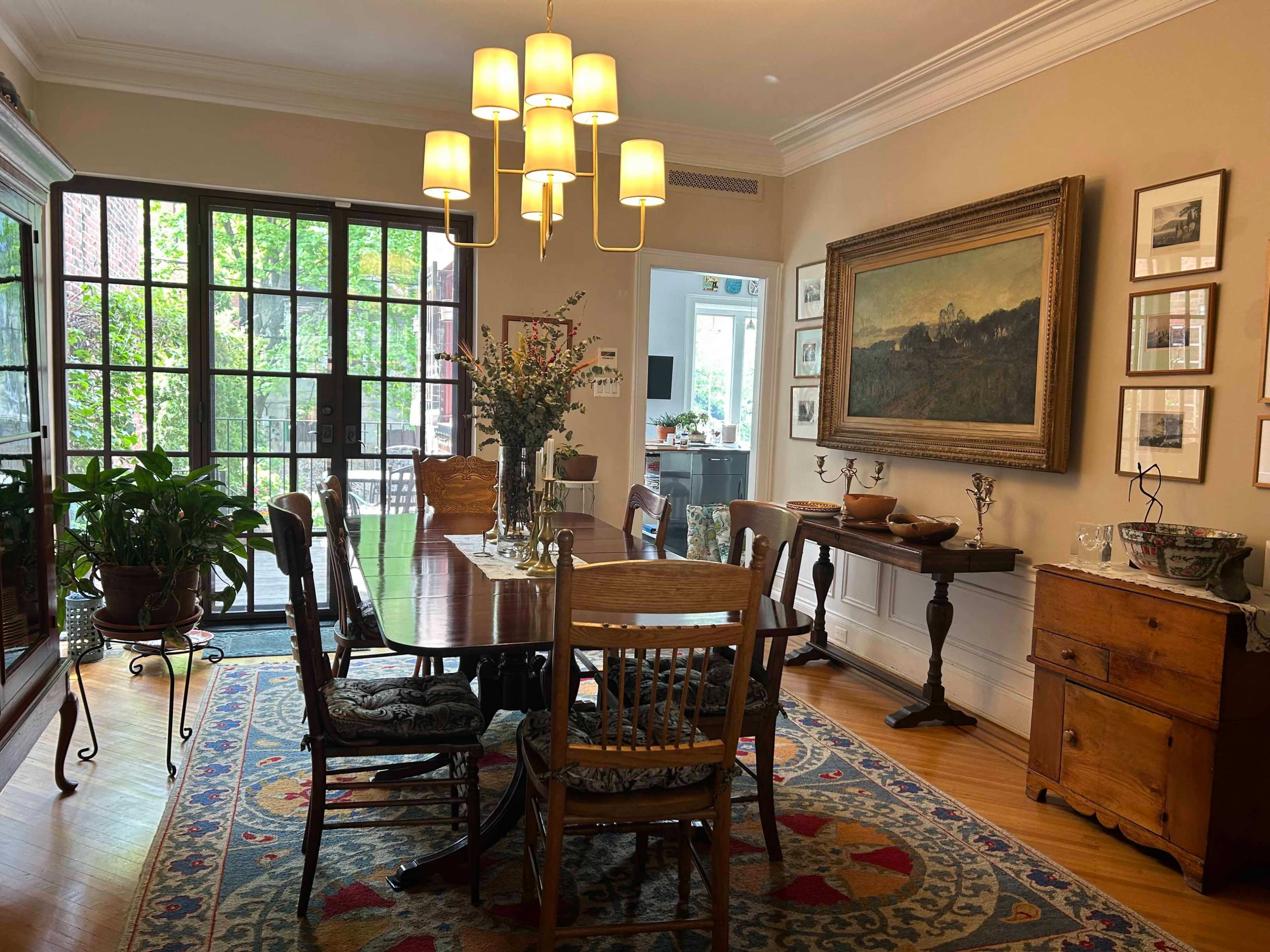 A dining room features a wooden table surrounded by chairs, a chandelier overhead, and artwork on the walls, with large windows letting in natural light.