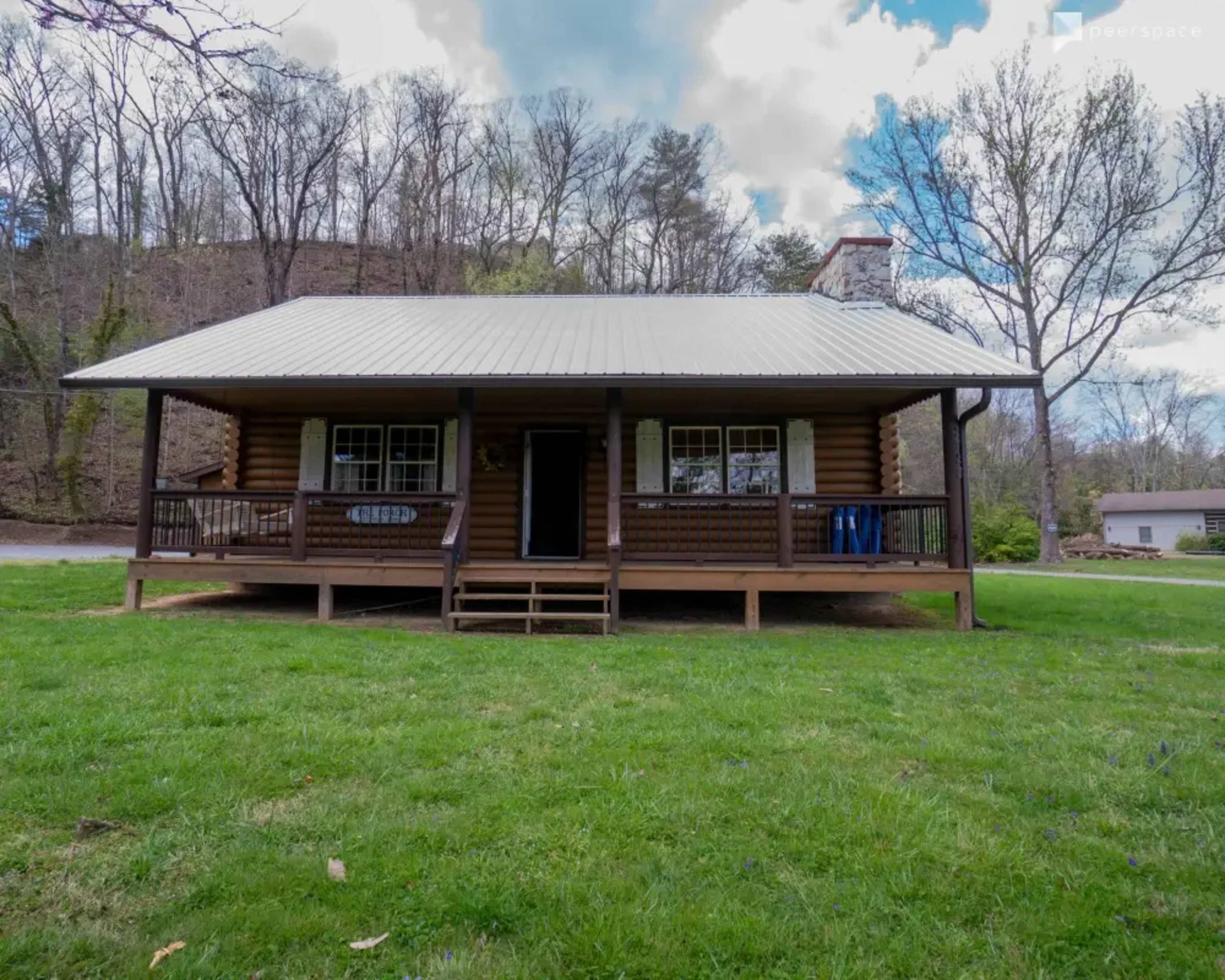 A wooden cabin with a metal roof and a front porch sits on a grassy area surrounded by trees under a cloudy sky.