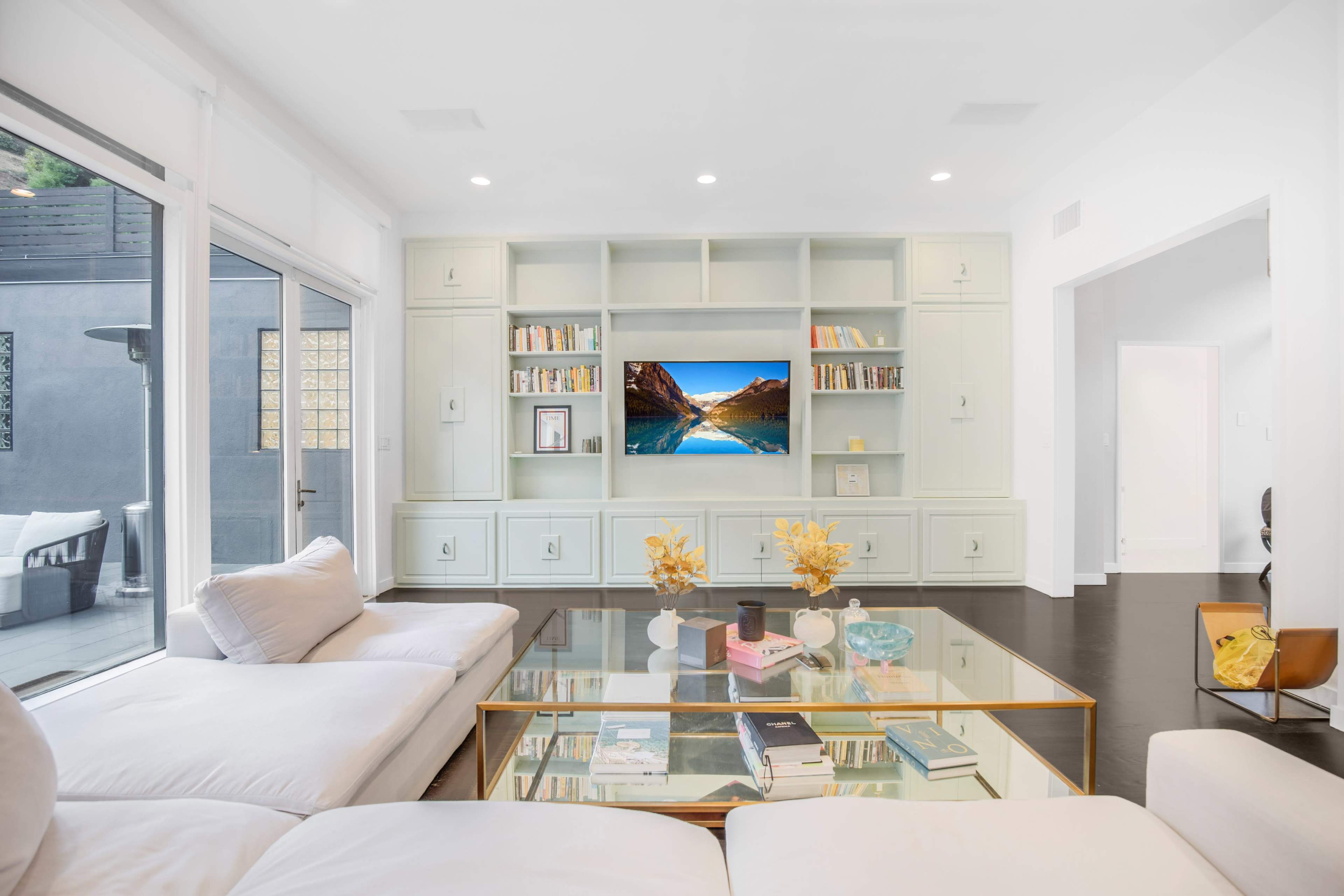 The living room features a light-colored sofa facing a modern built-in shelf with a TV and books, and a glass coffee table with decorative items.