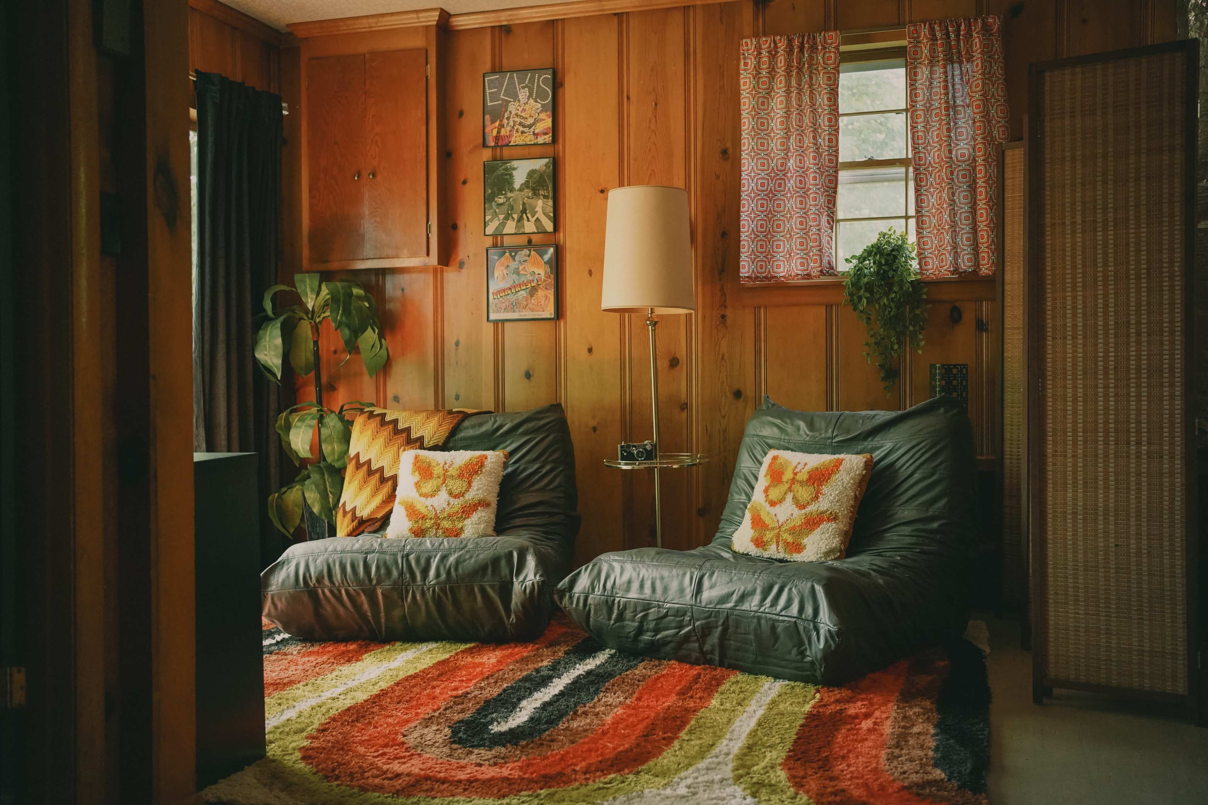 A living room features two green lounge chairs, a colorful patterned rug, and wooden paneled walls with framed artwork.
