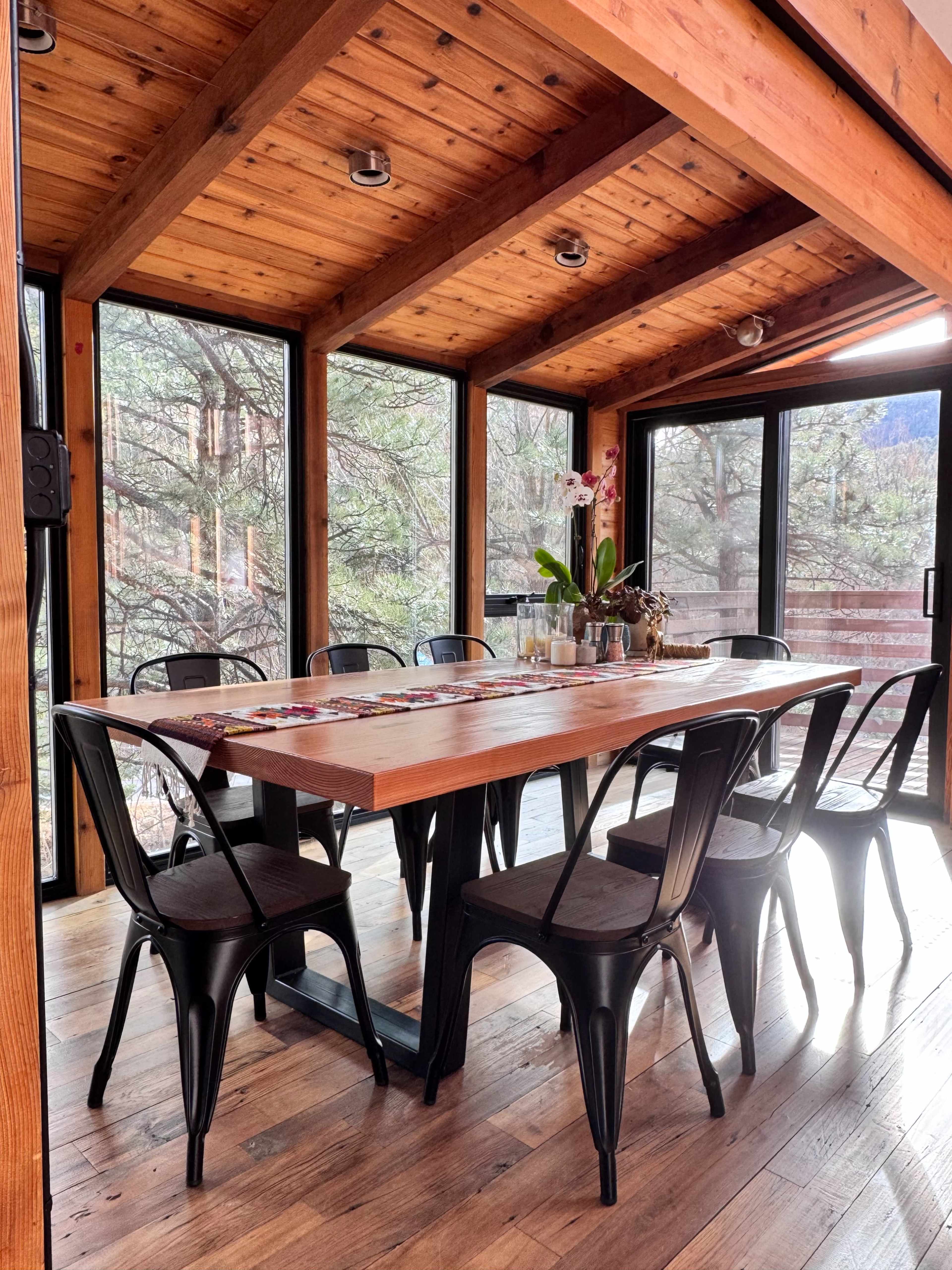 A wooden dining table with metal chairs is set in a sunlit room surrounded by large windows, showcasing views of trees outside.