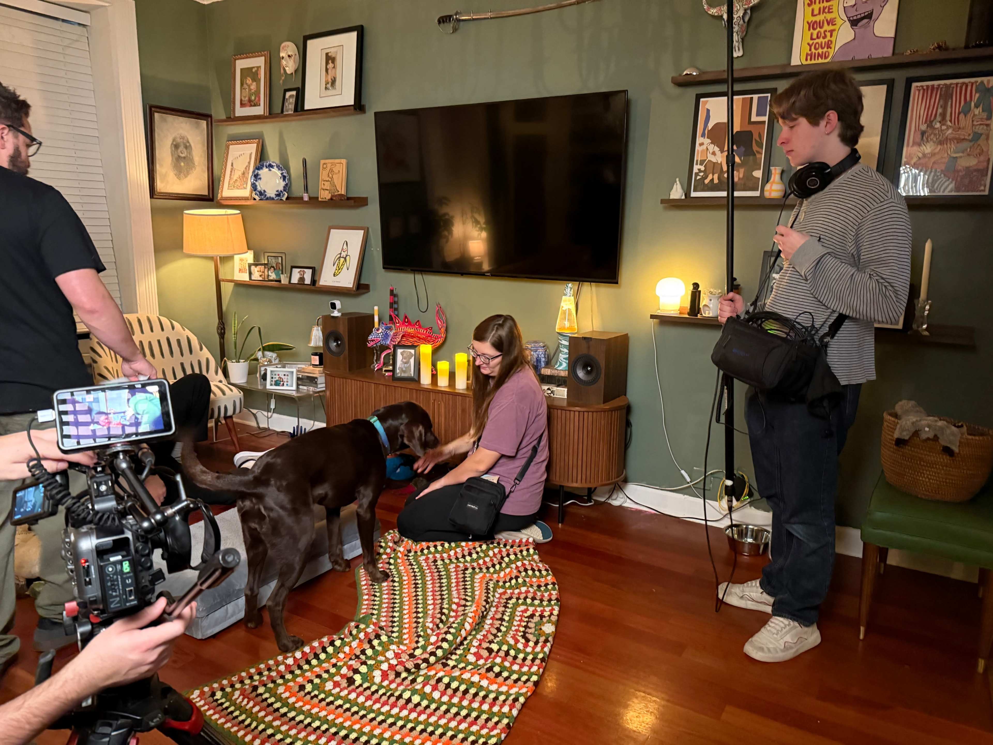 A crew is filming in a living room with a television, where a woman interacts with a chocolate lab on a colorful blanket while another person holds audio equipment.