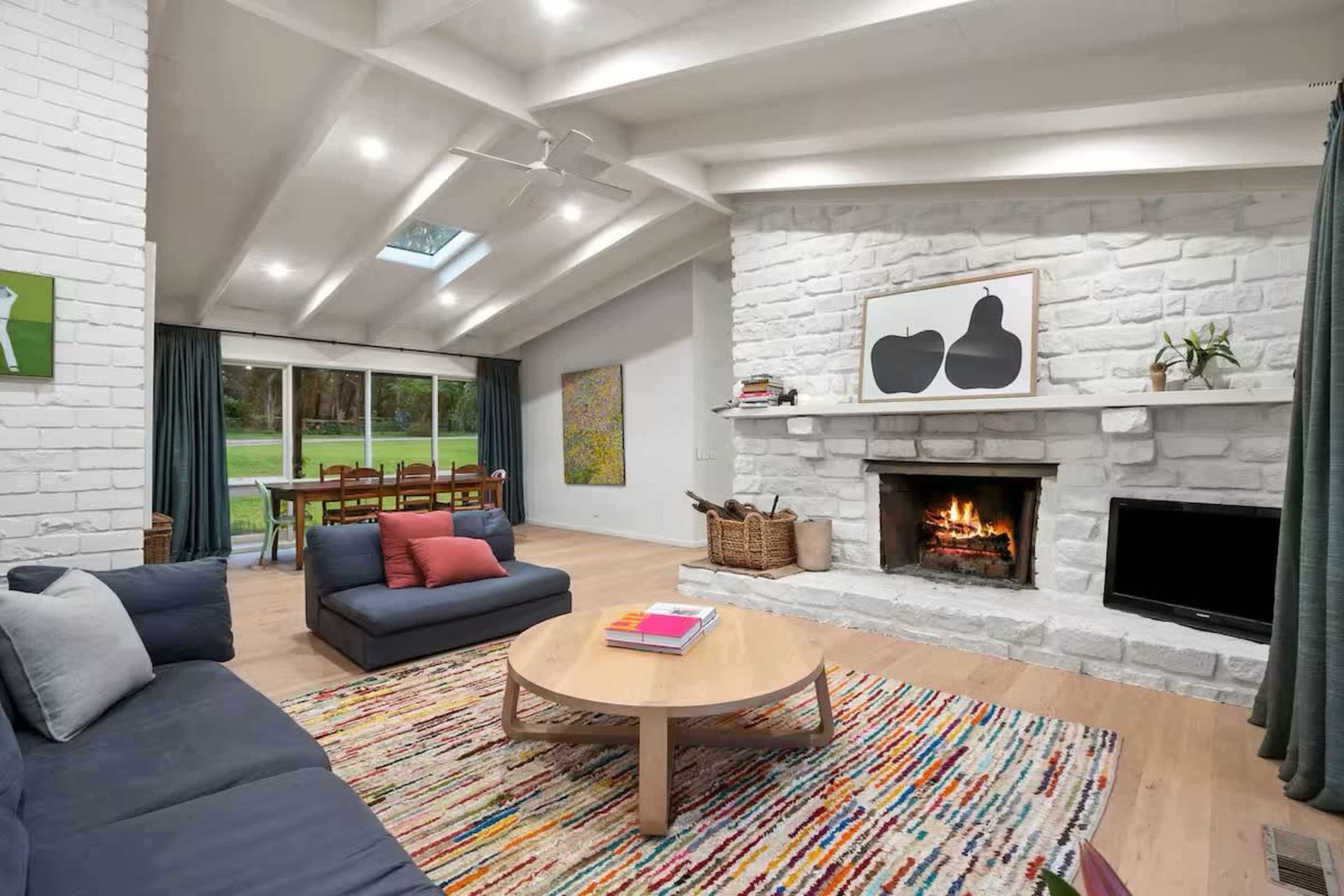 A modern living room features a stone fireplace, large windows with a view of a dining area, and a rug with colorful stripes.