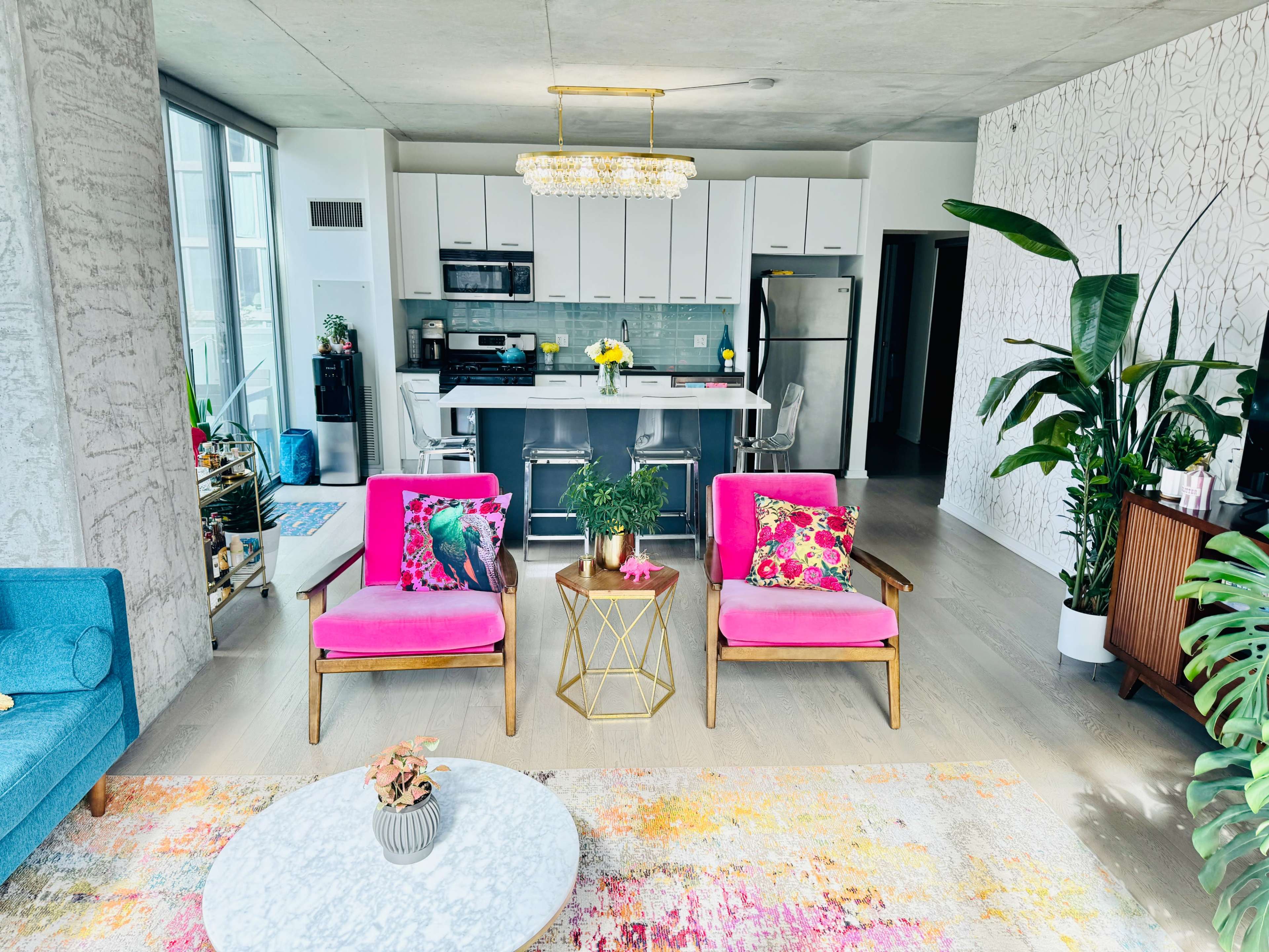A bright, modern living room with a kitchen in the background, featuring two pink chairs with patterned pillows, a coffee table, and decorative plants.