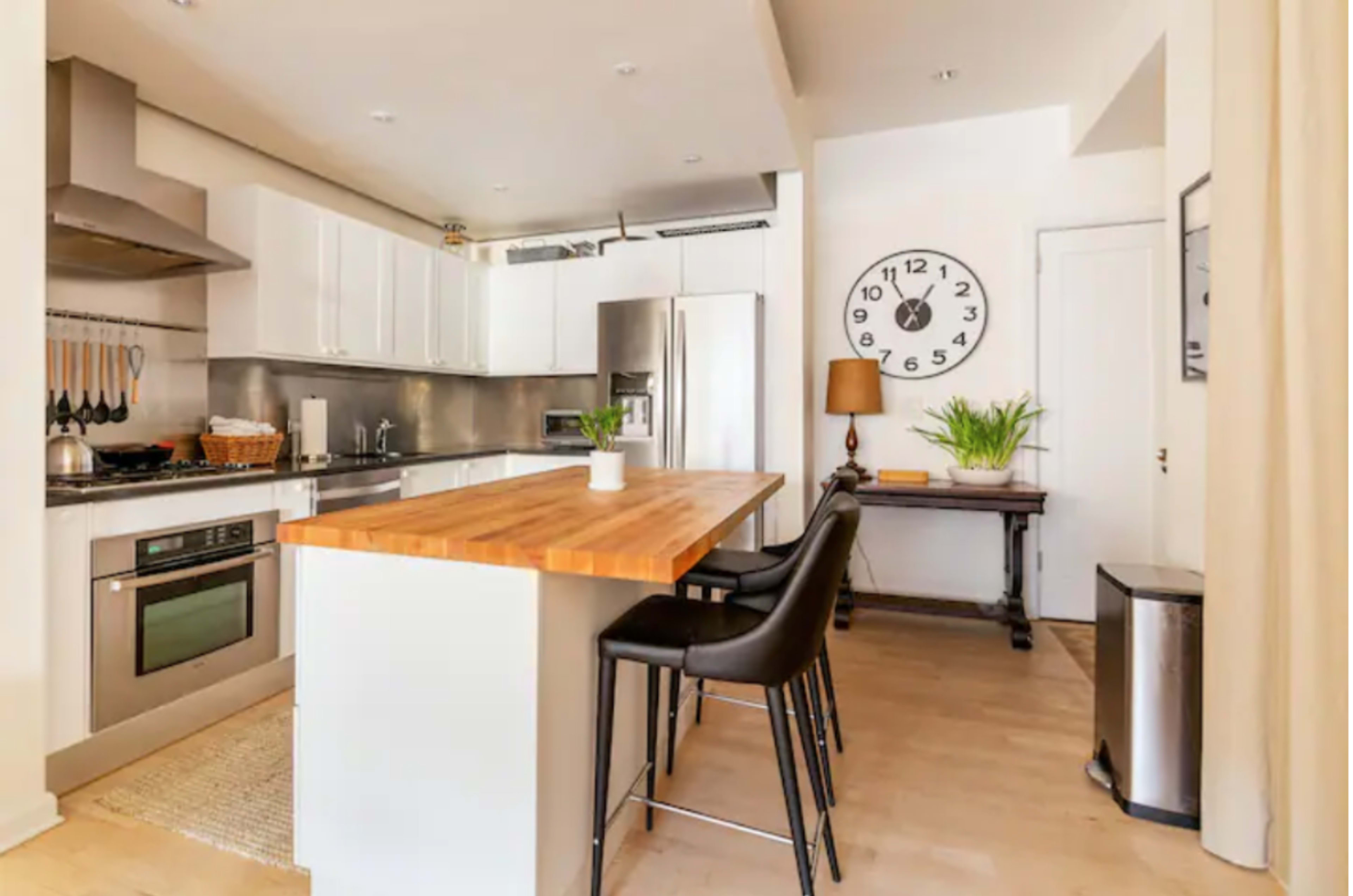 A modern kitchen with a wooden island, bar stools, stainless steel appliances, and a large wall clock.