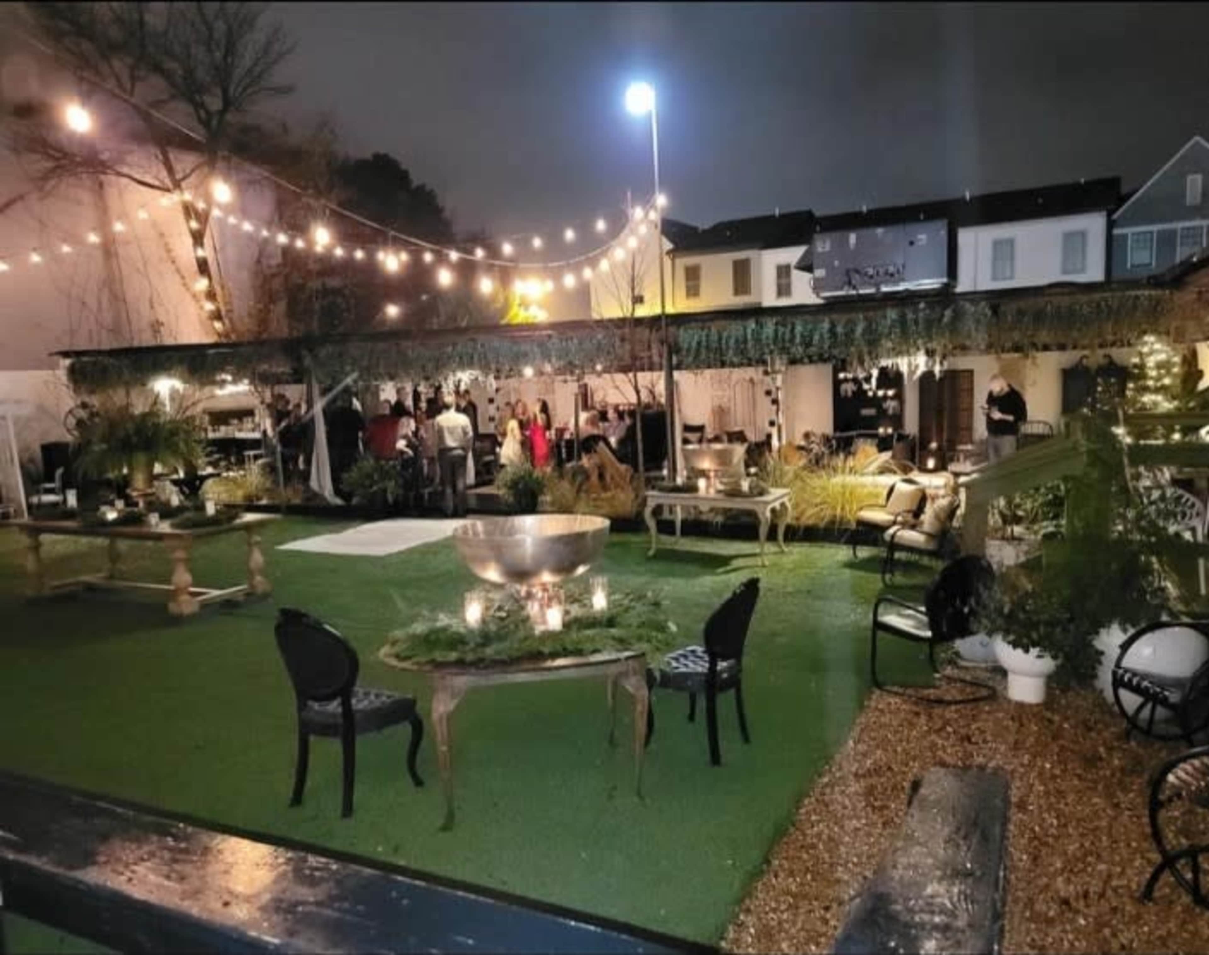 An outdoor event space with string lights, tables, and seating arranged on a lawn, creating a festive atmosphere.