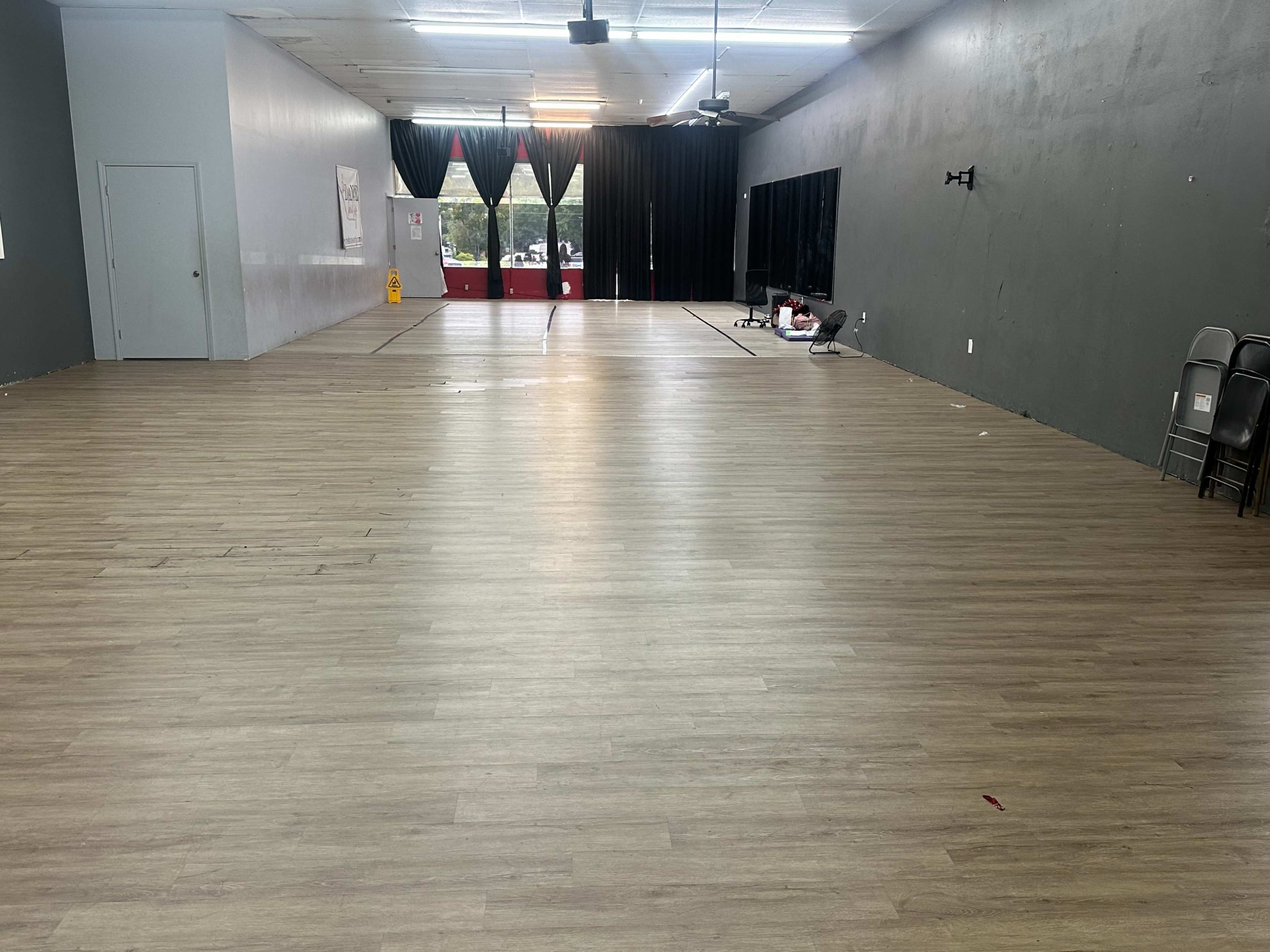 The image shows an empty indoor space with wooden flooring and gray walls, featuring a stage area and large windows covered by black curtains.