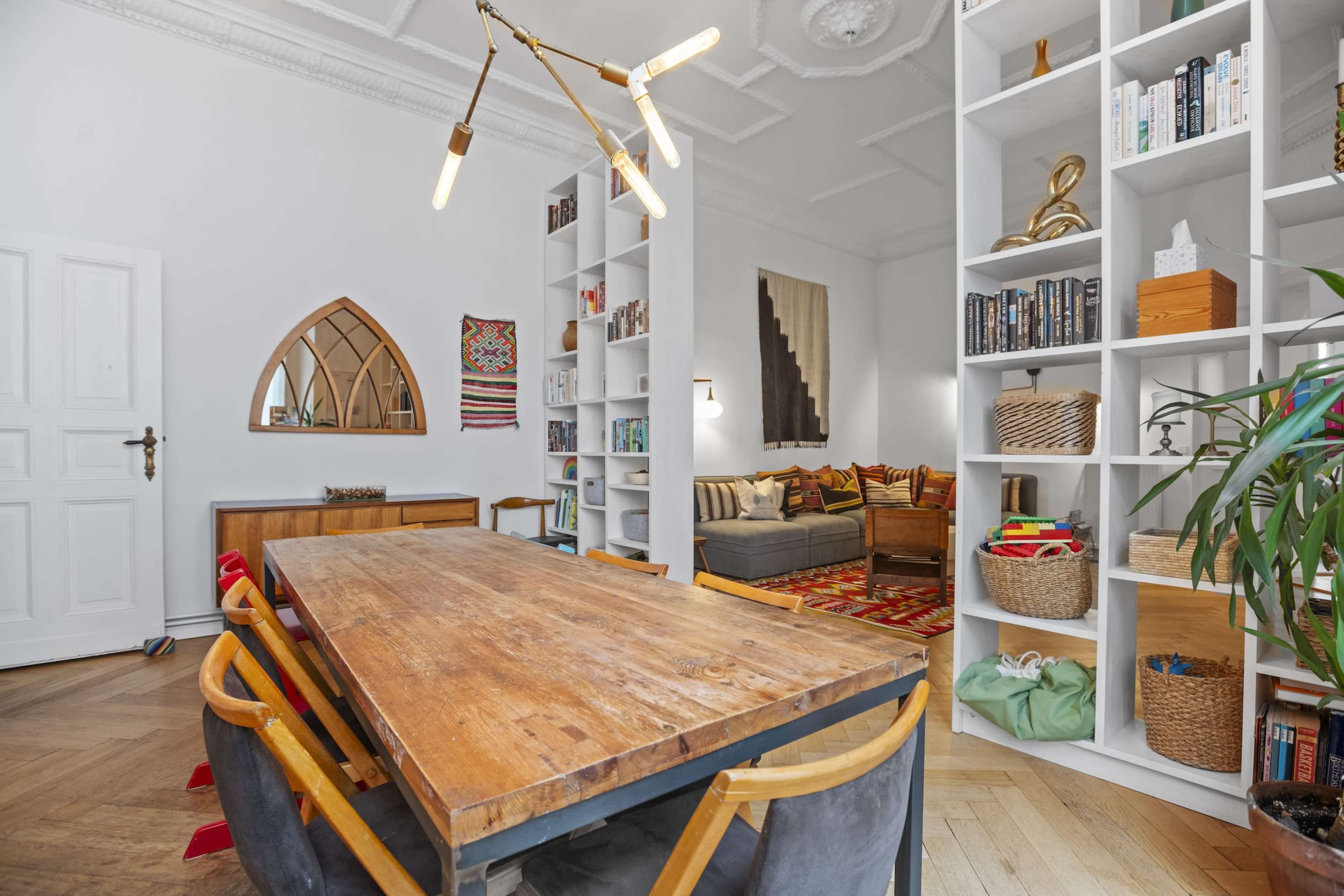The image shows a modern dining area featuring a wooden table and chairs, with bookshelves and decorative items in a well-lit, stylish room.