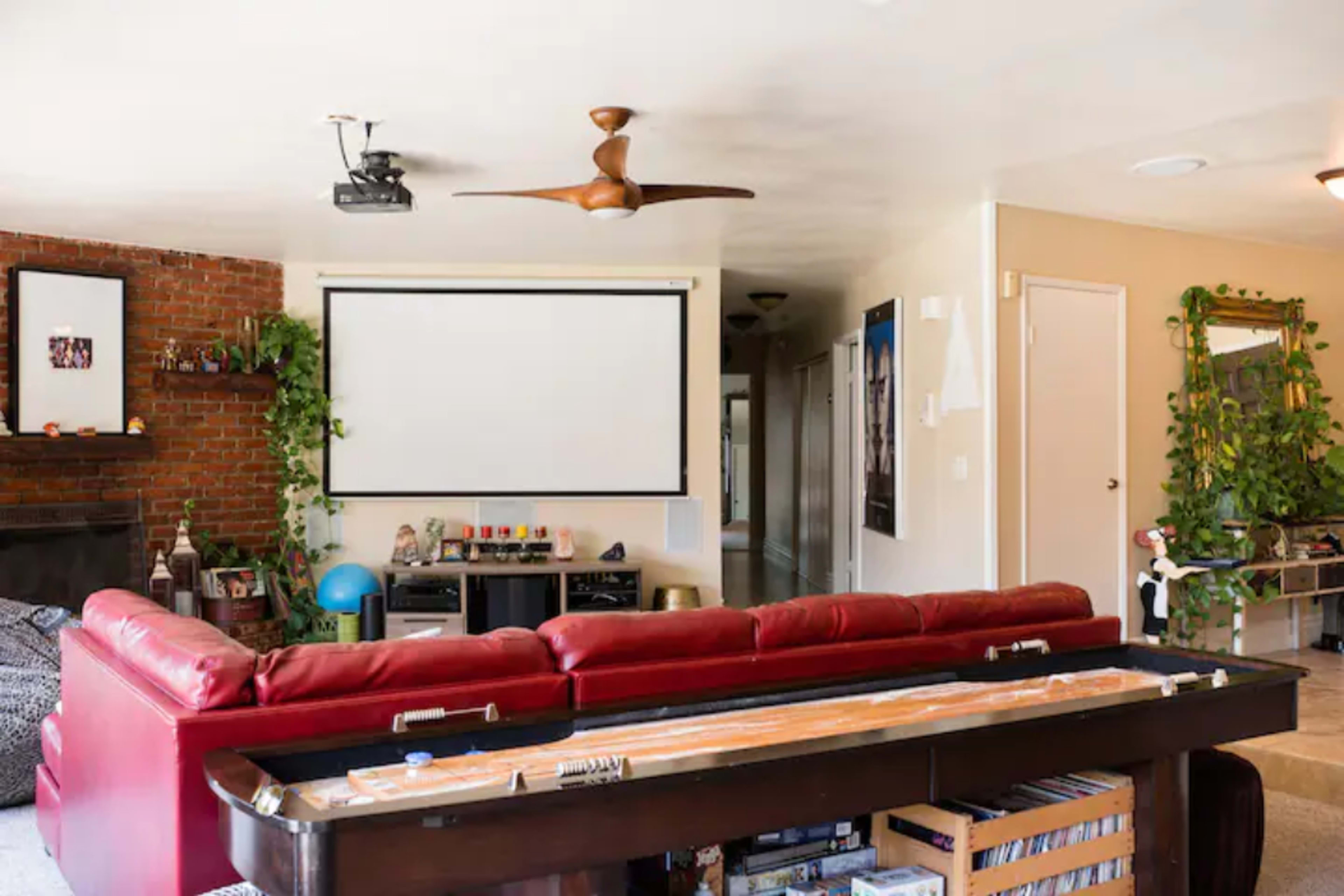 A living room with a red sectional sofa, a large projector screen mounted on the wall, and a pool table in the foreground.