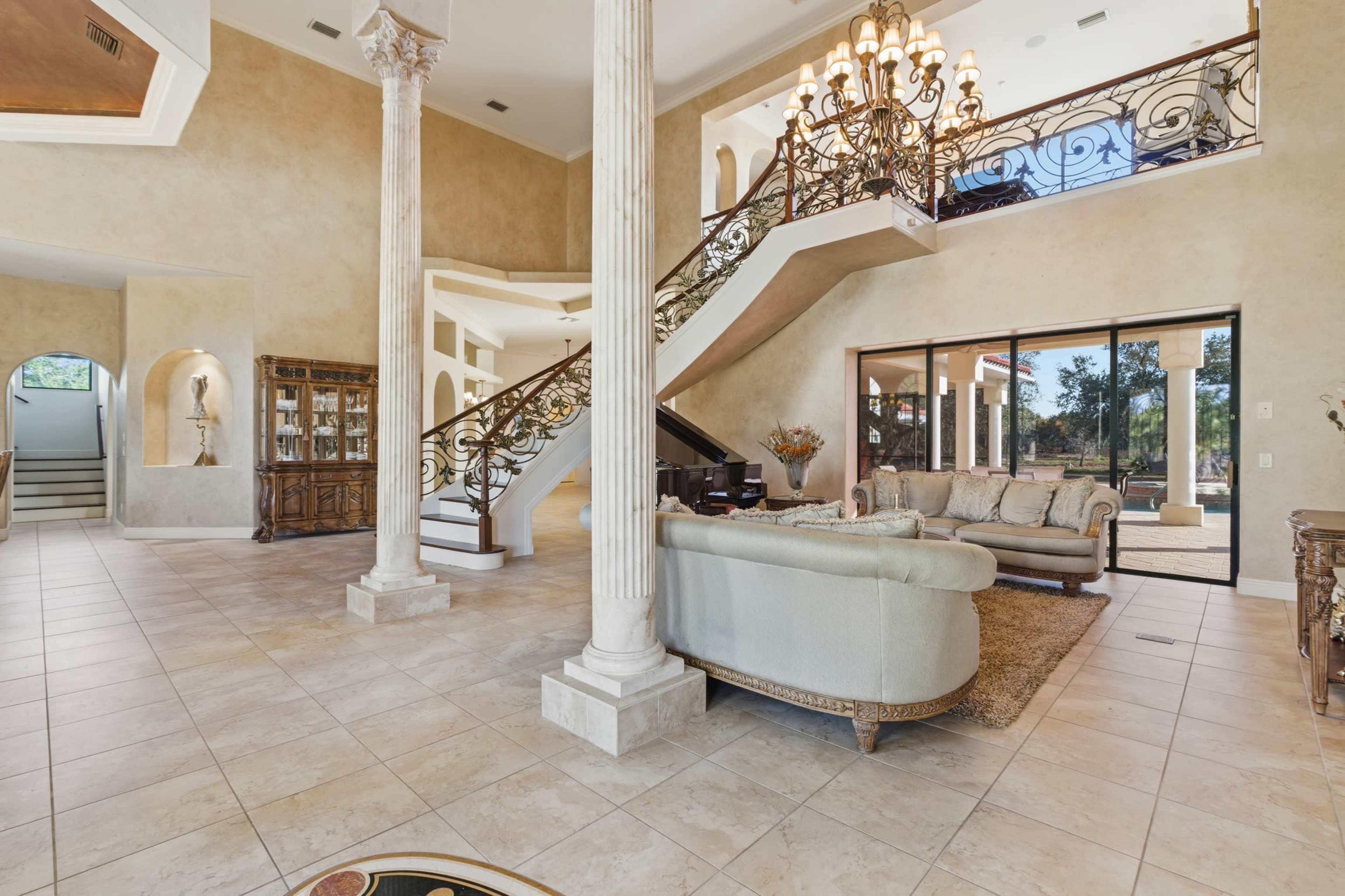 The image shows a spacious entryway with a grand staircase, decorative columns, and a living area featuring a large sofa and a glass wall overlooking a garden.