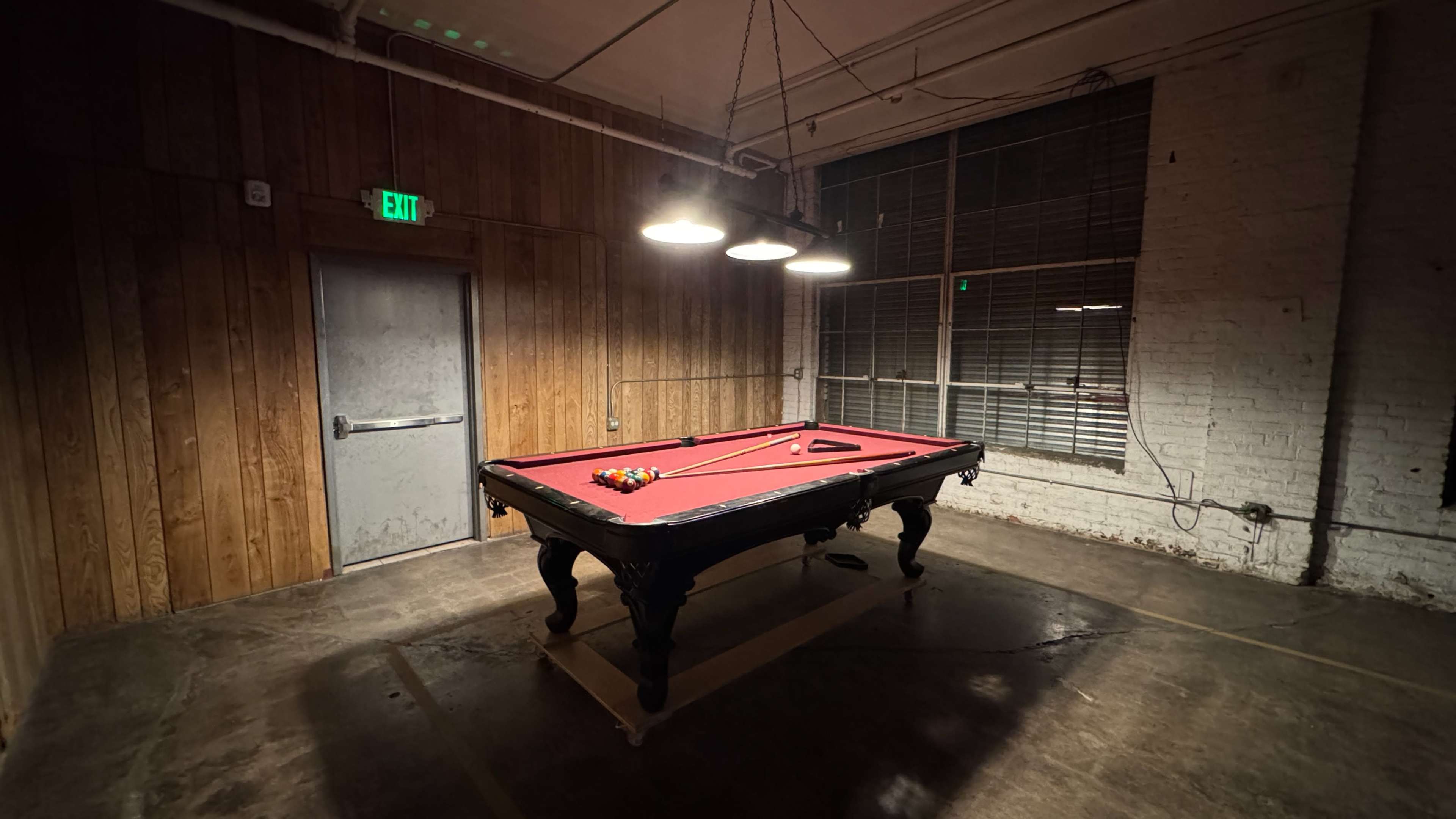 A red pool table with a set of balls rests in a dimly lit room with wooden walls and a large window.