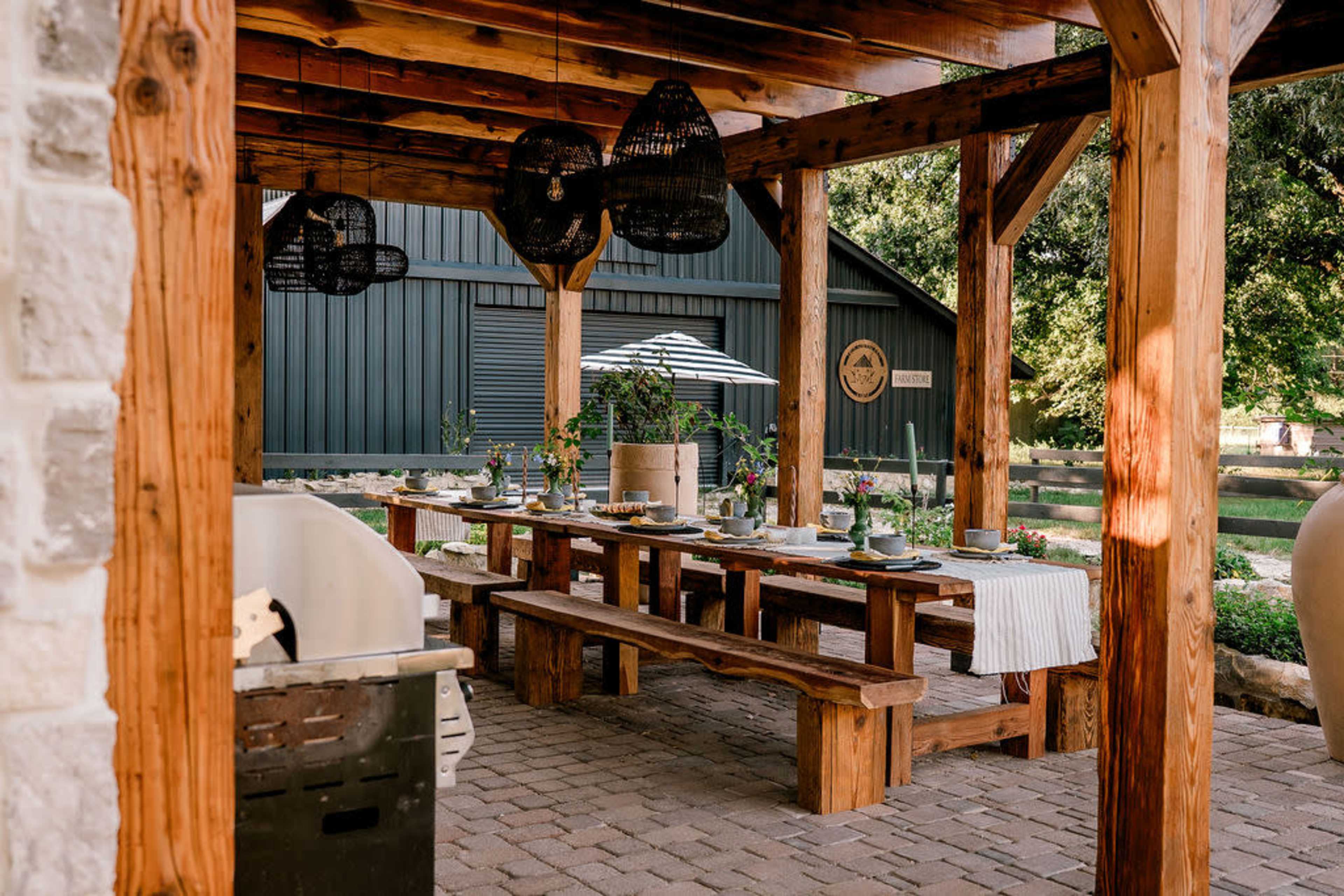 The image shows a rustic outdoor dining area with wooden tables, benches, and hanging light fixtures under a covered structure.