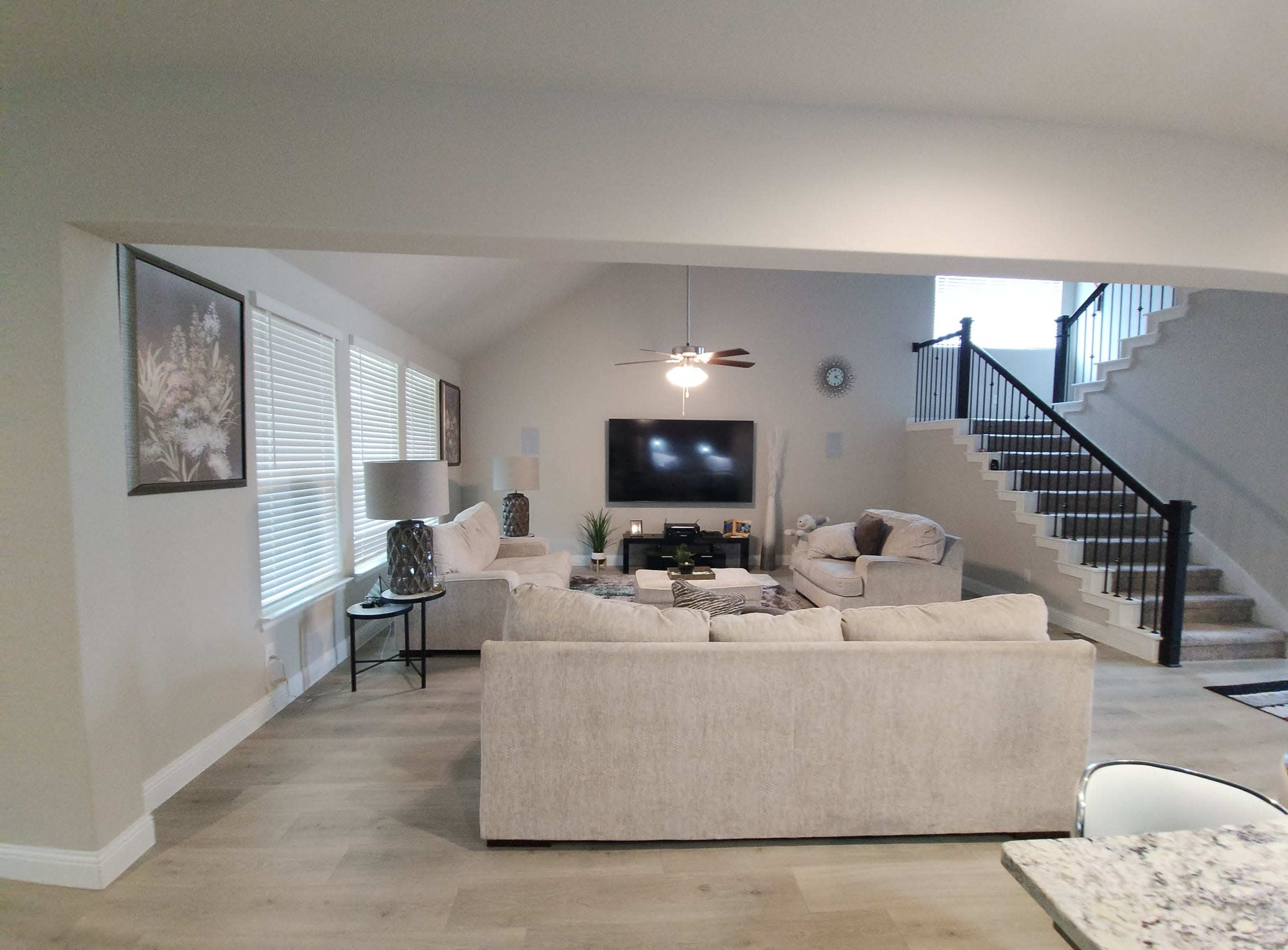 A spacious living room with two light-colored sofas facing a wall-mounted television, adjacent to a staircase leading to an upper level.