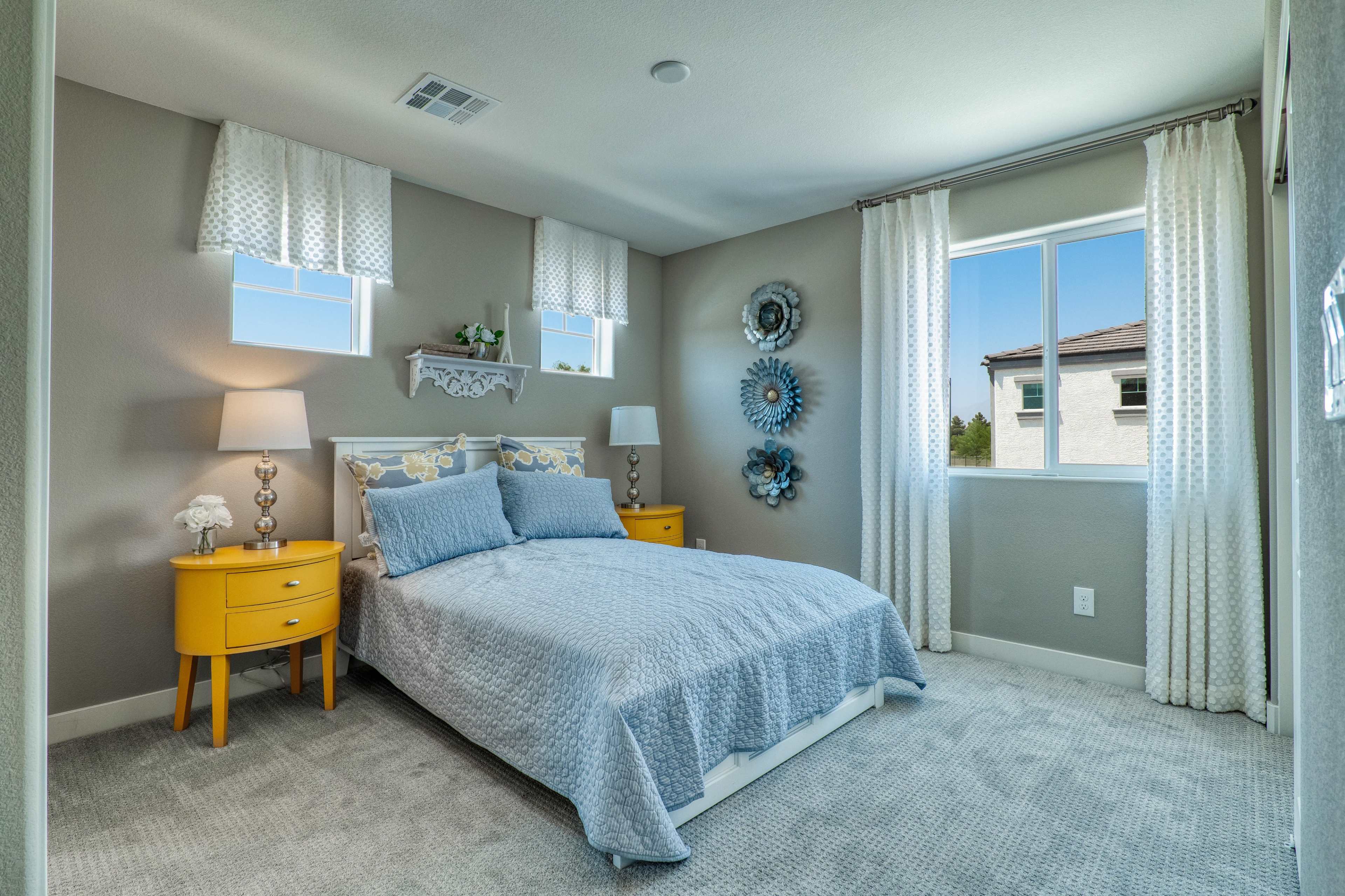 A neatly arranged bedroom features a bed with a light blue coverlet, two bedside tables, and wall decor including circular mirrors and window treatments.