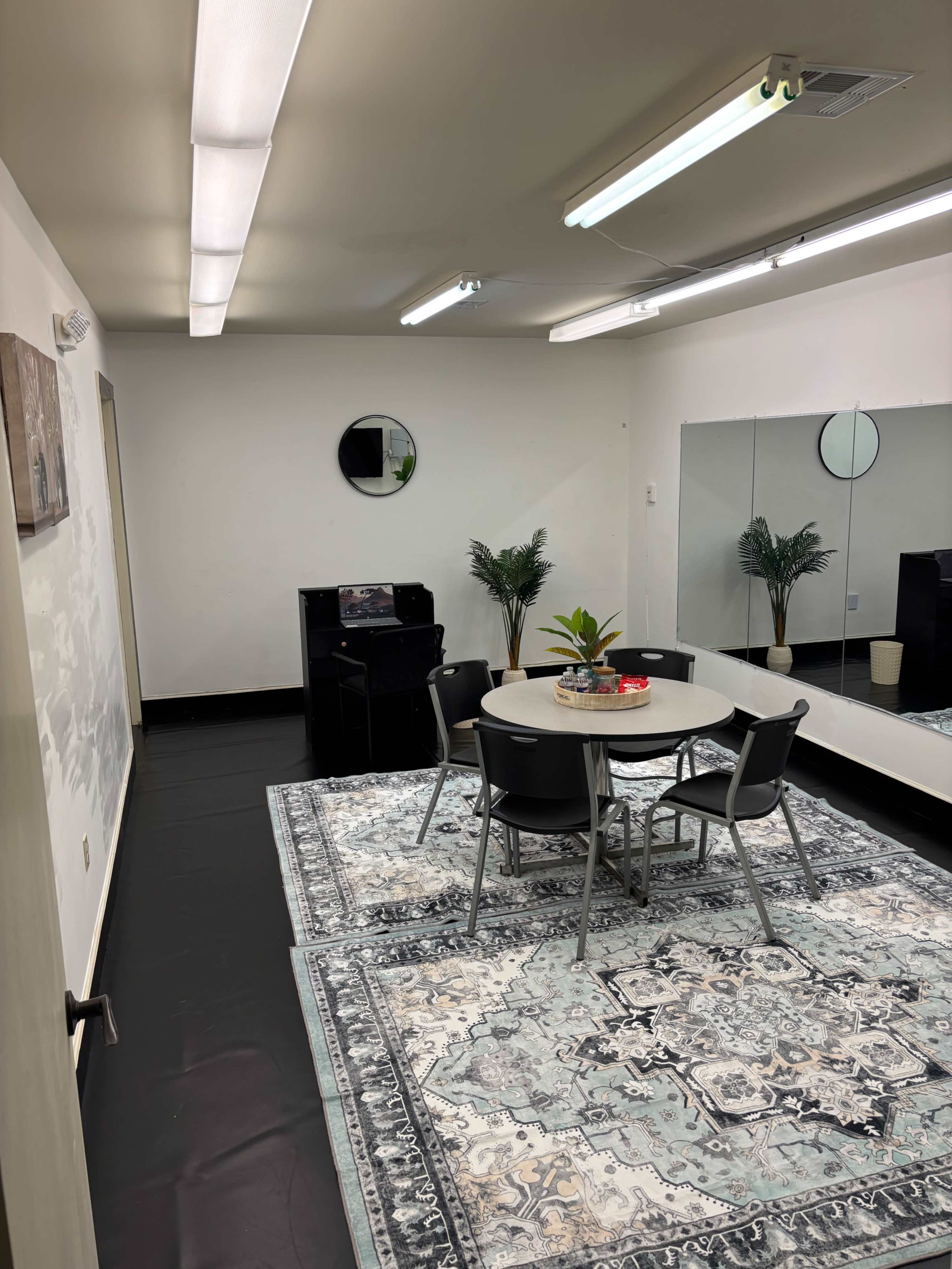 The image shows a simple interior room with a round table and four chairs on a patterned rug, flanked by two plants, and featuring mirrored walls and a small cabinet.