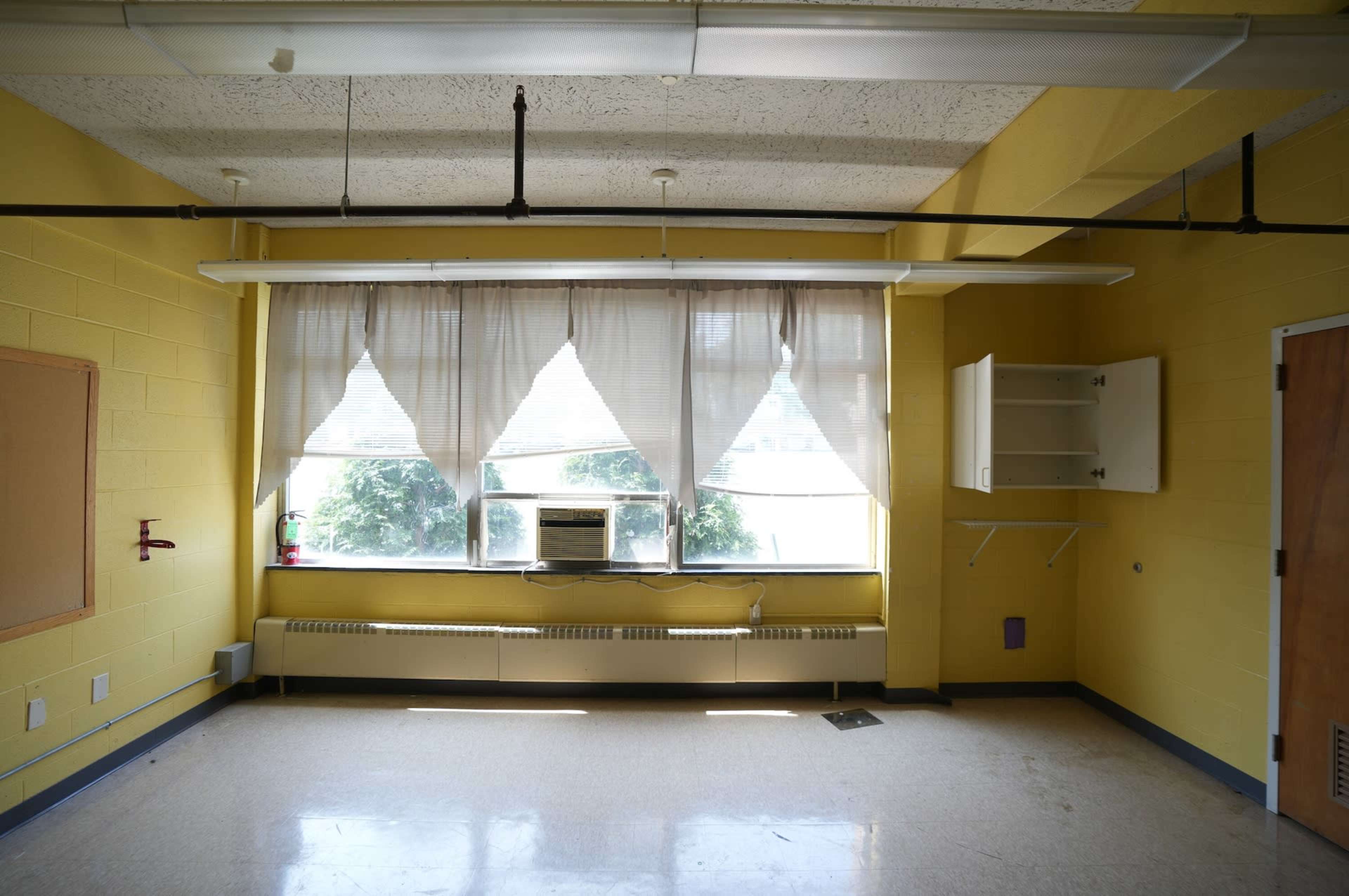 The image shows a brightly colored yellow room with large windows covered by sheer curtains, featuring an air conditioning unit and minimal furnishings.