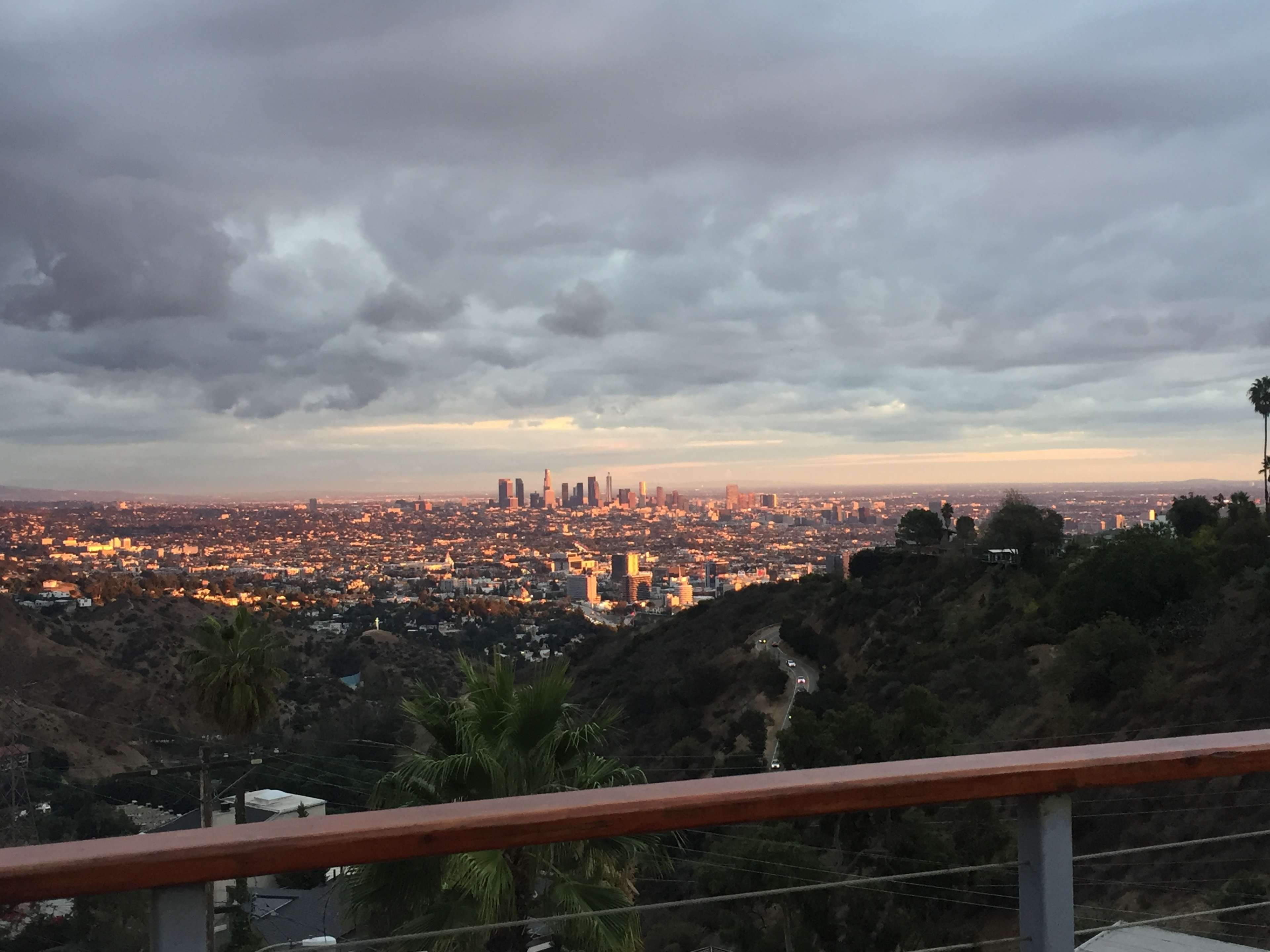 The image shows a panoramic view of a city at dusk, with buildings illuminated against a cloudy sky and hills in the foreground.