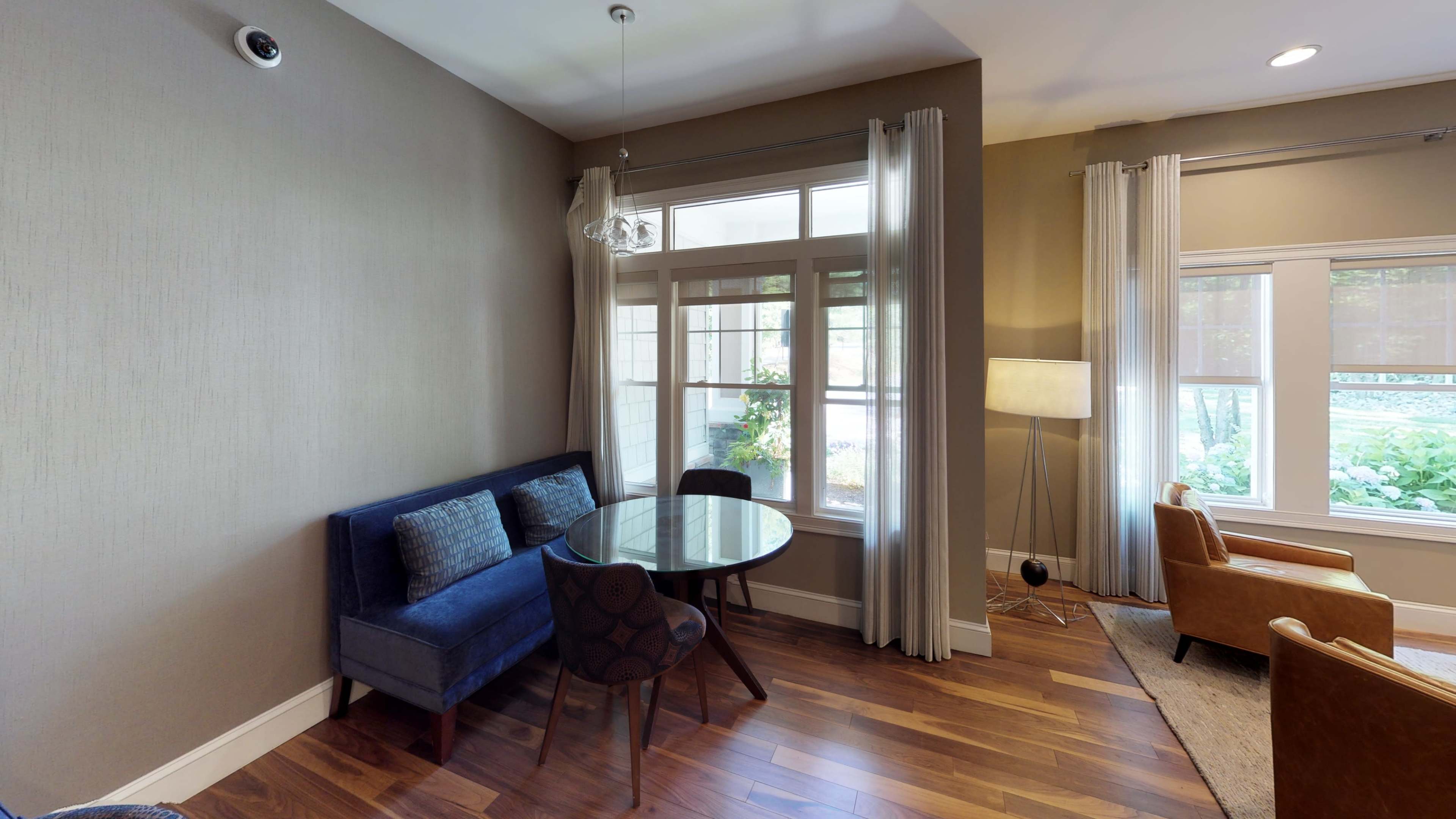 A small sitting area featuring a blue couch, a round glass table, and two wooden chairs with a window providing natural light.