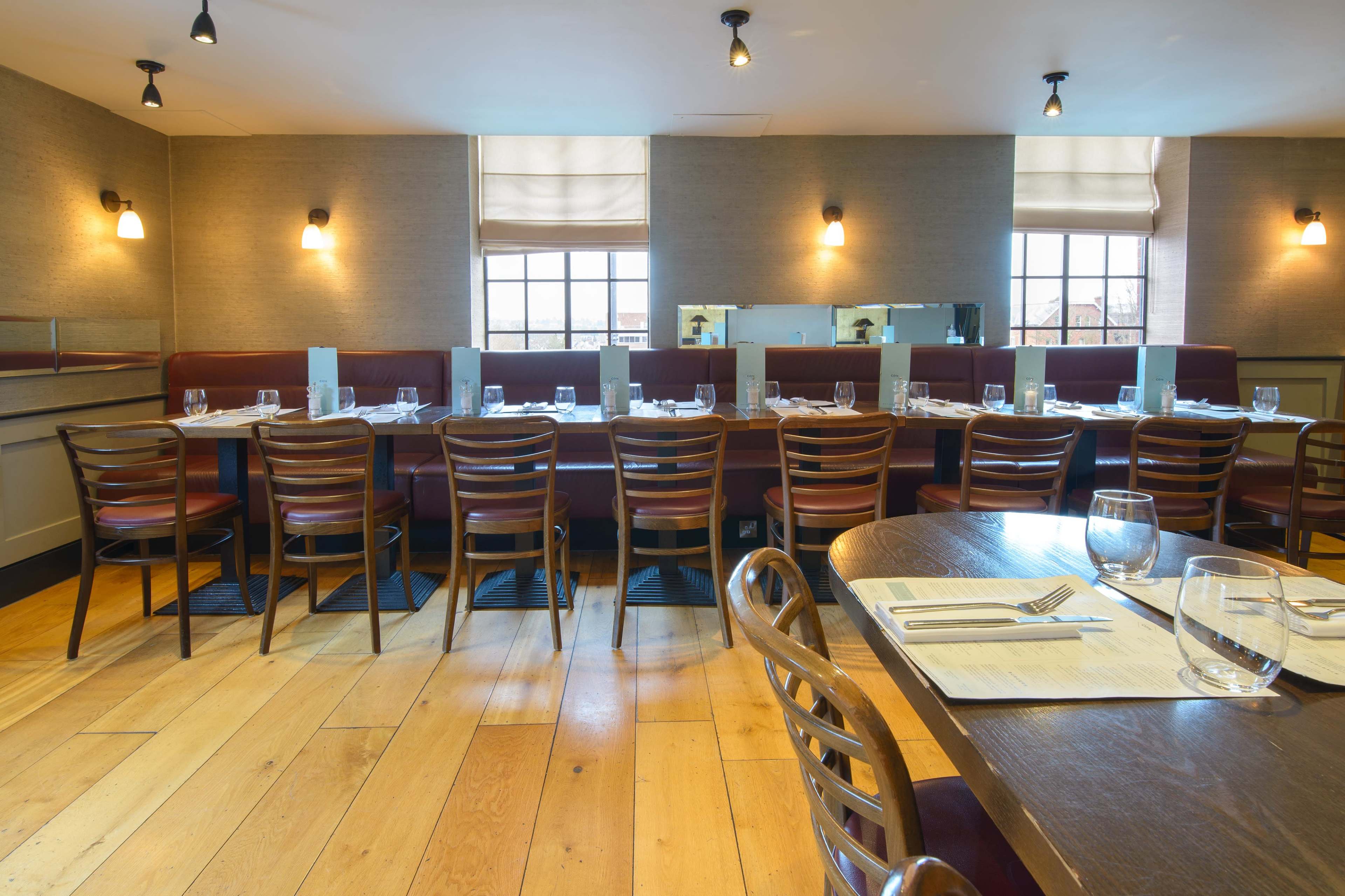 The image shows an elegantly arranged restaurant interior with wooden flooring, tables set with glassware, and a row of chairs lining a central table.
