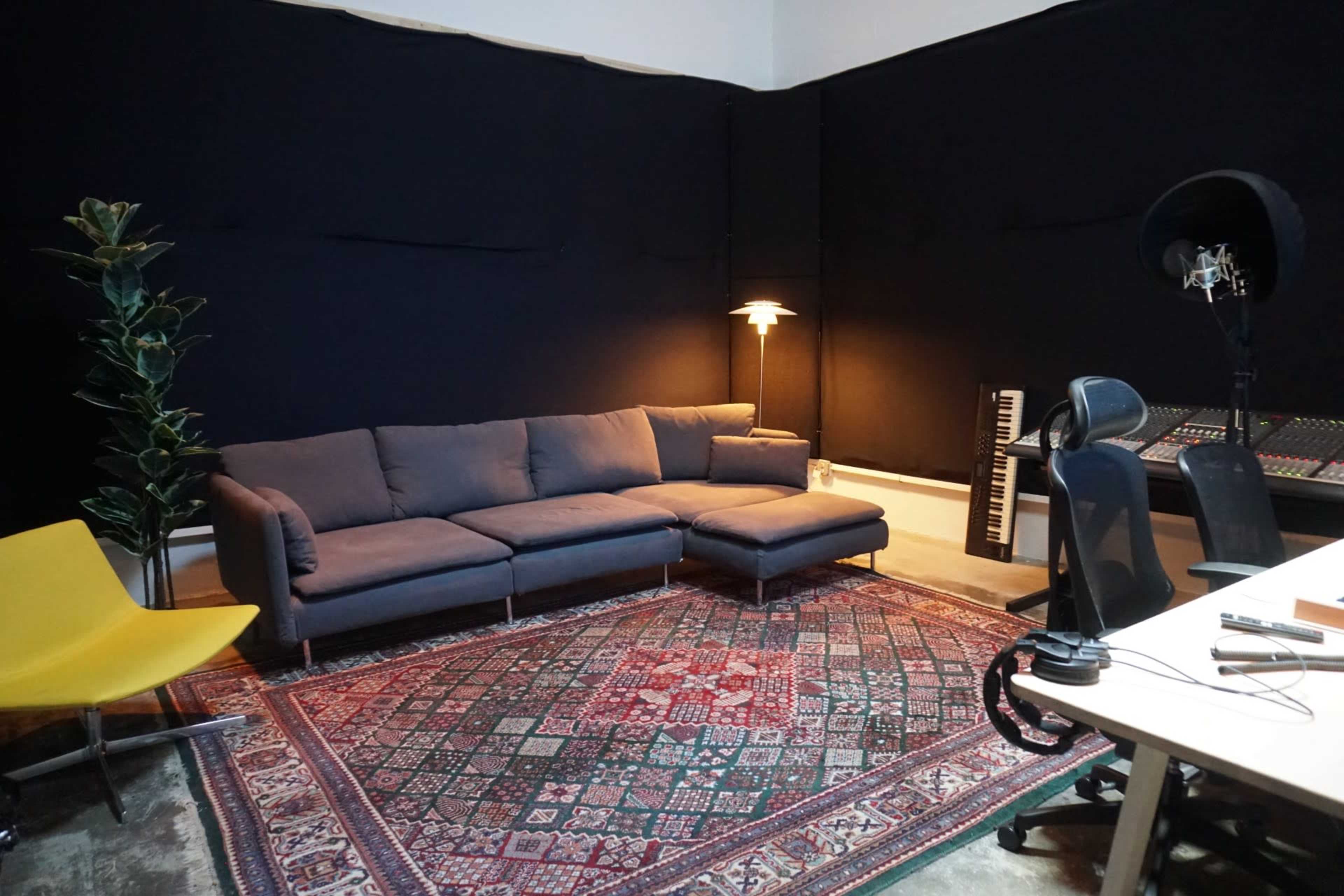 The image shows a studio space featuring a gray sectional couch, a floor lamp, a keyboard, and an area rug, with a mixing console and office chairs nearby.