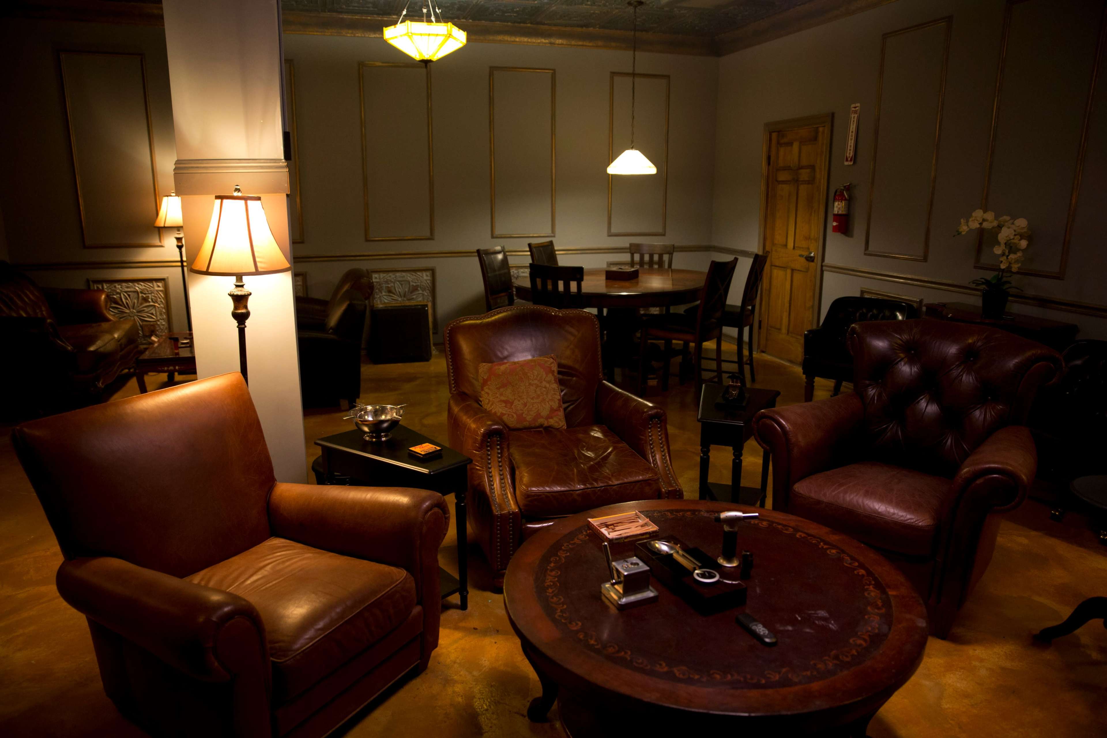 A dimly lit lounge area with brown leather armchairs, a round table, and a dining setup in the background.