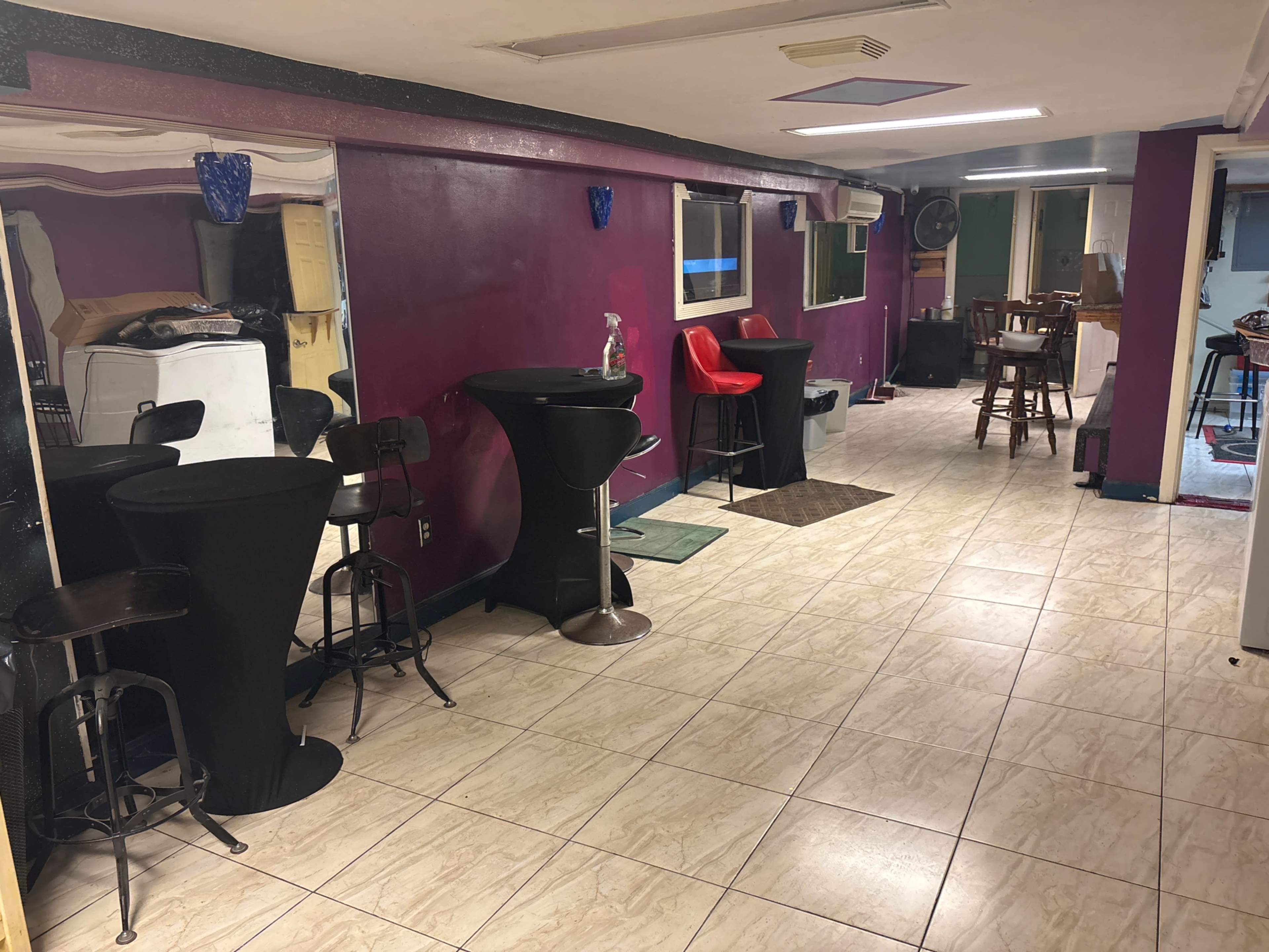 The image shows a narrow room with tiled flooring featuring bar-height tables and chairs, a red chair, and multiple windows providing natural light.