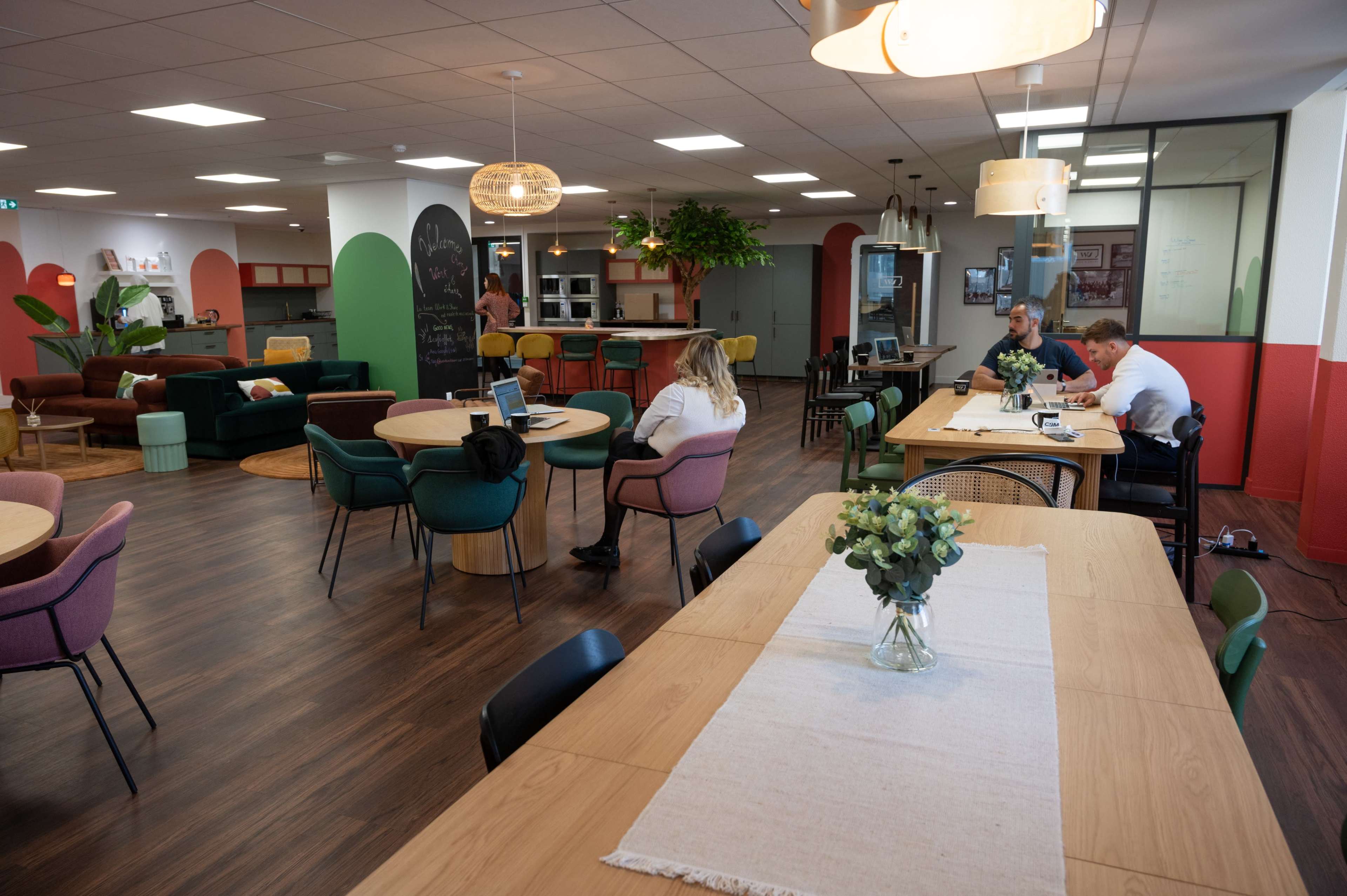 A modern open office space features various seating areas, wooden tables, and plants, with people engaged in conversation and work.