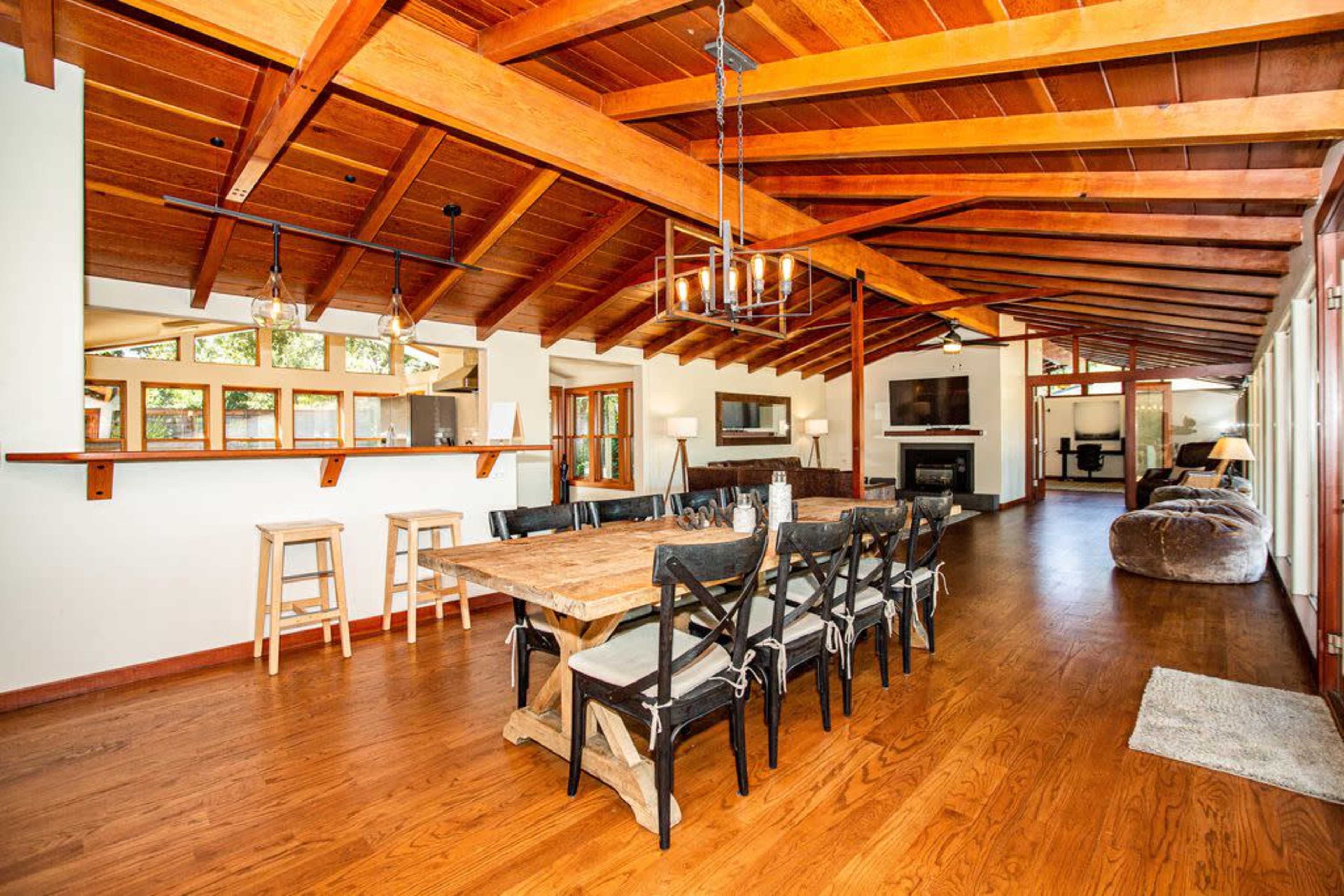 The image shows a spacious wooden dining area with a long table and black chairs, leading to a well-lit living space featuring a fireplace and a bar counter.