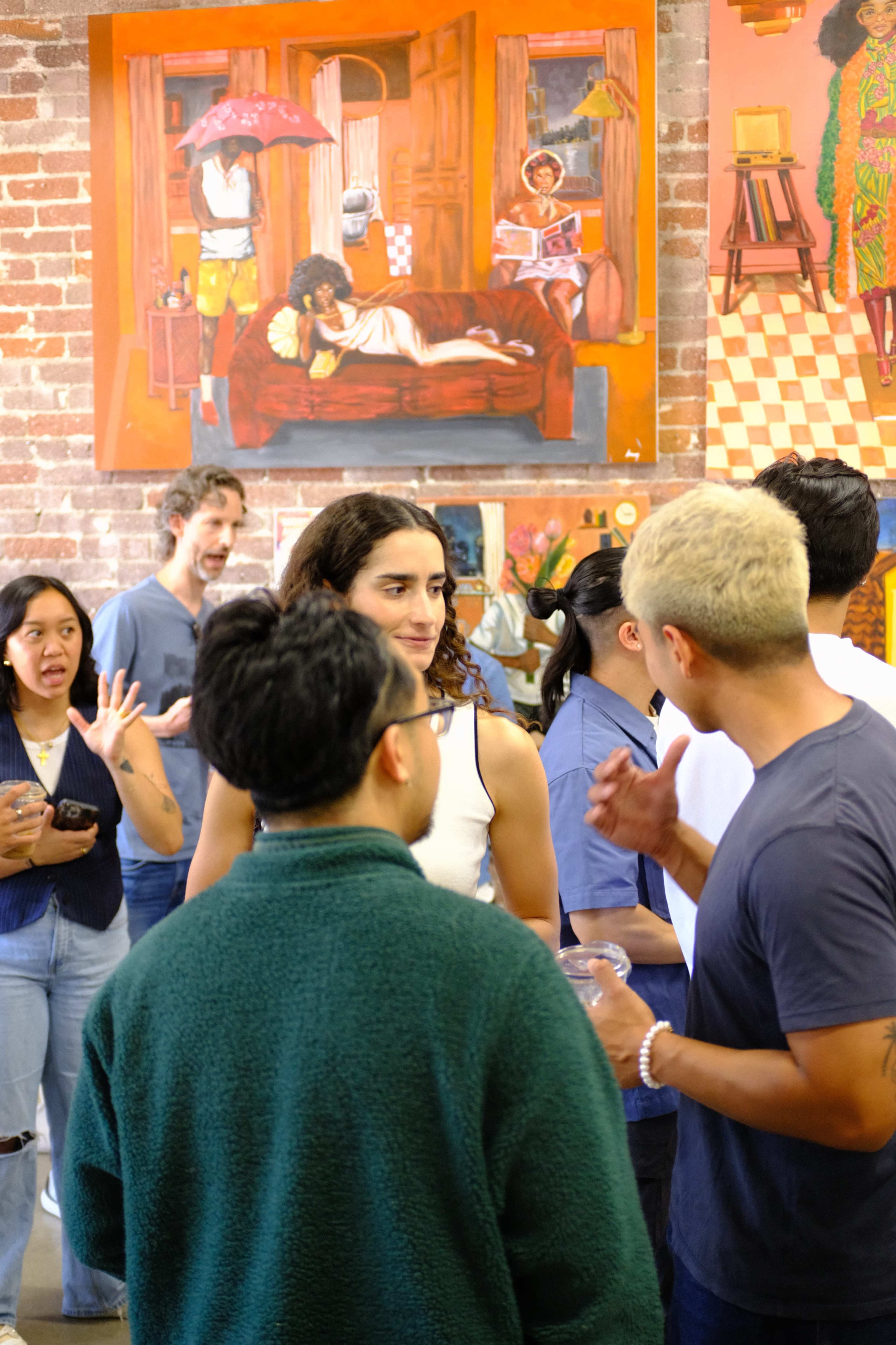 A group of people is engaging in conversation during an event in an art space, surrounded by vibrant paintings on the walls.