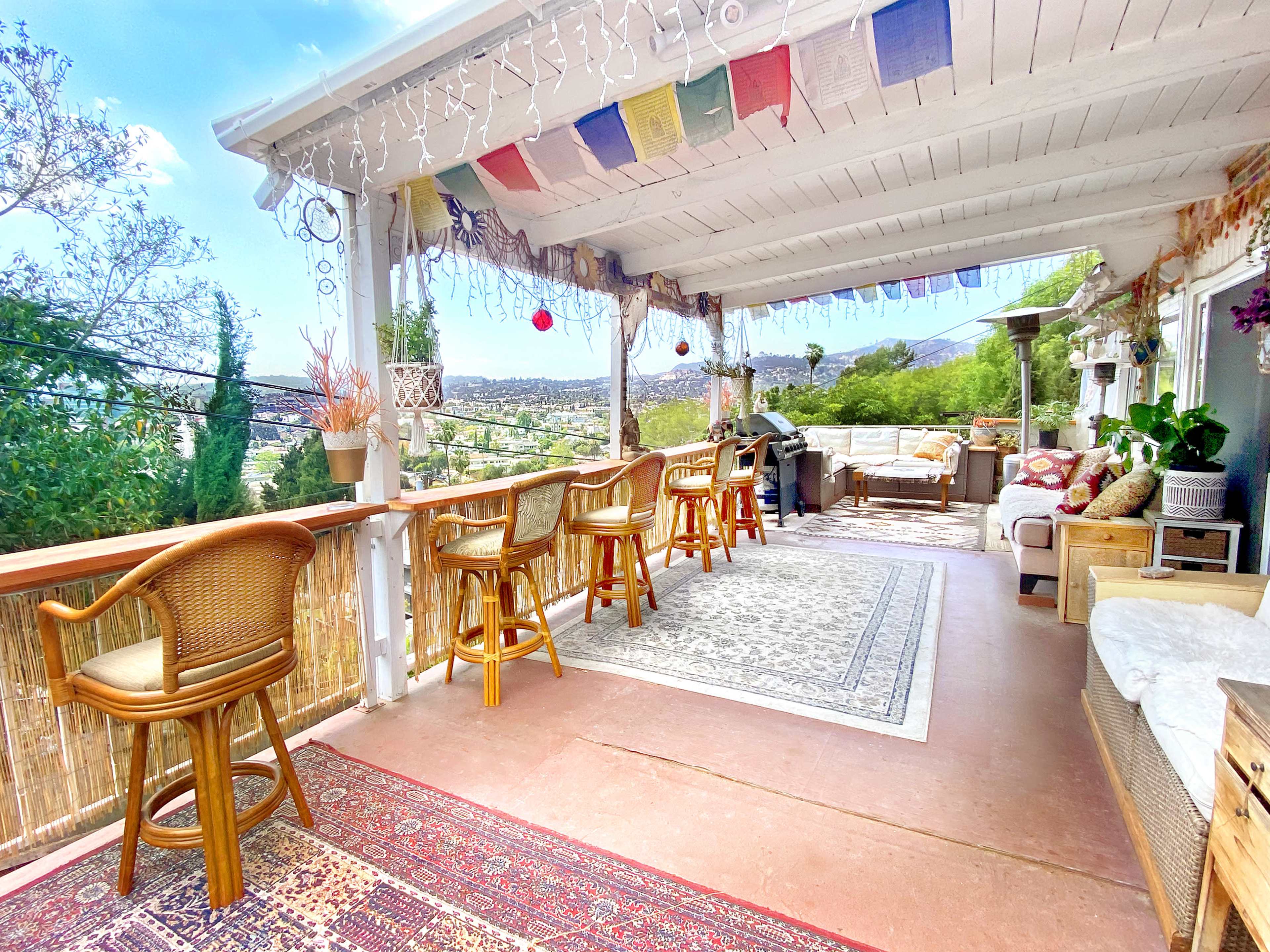A spacious outdoor patio with a bar area, chairs, rugs, and decorative plants overlooking a scenic landscape.