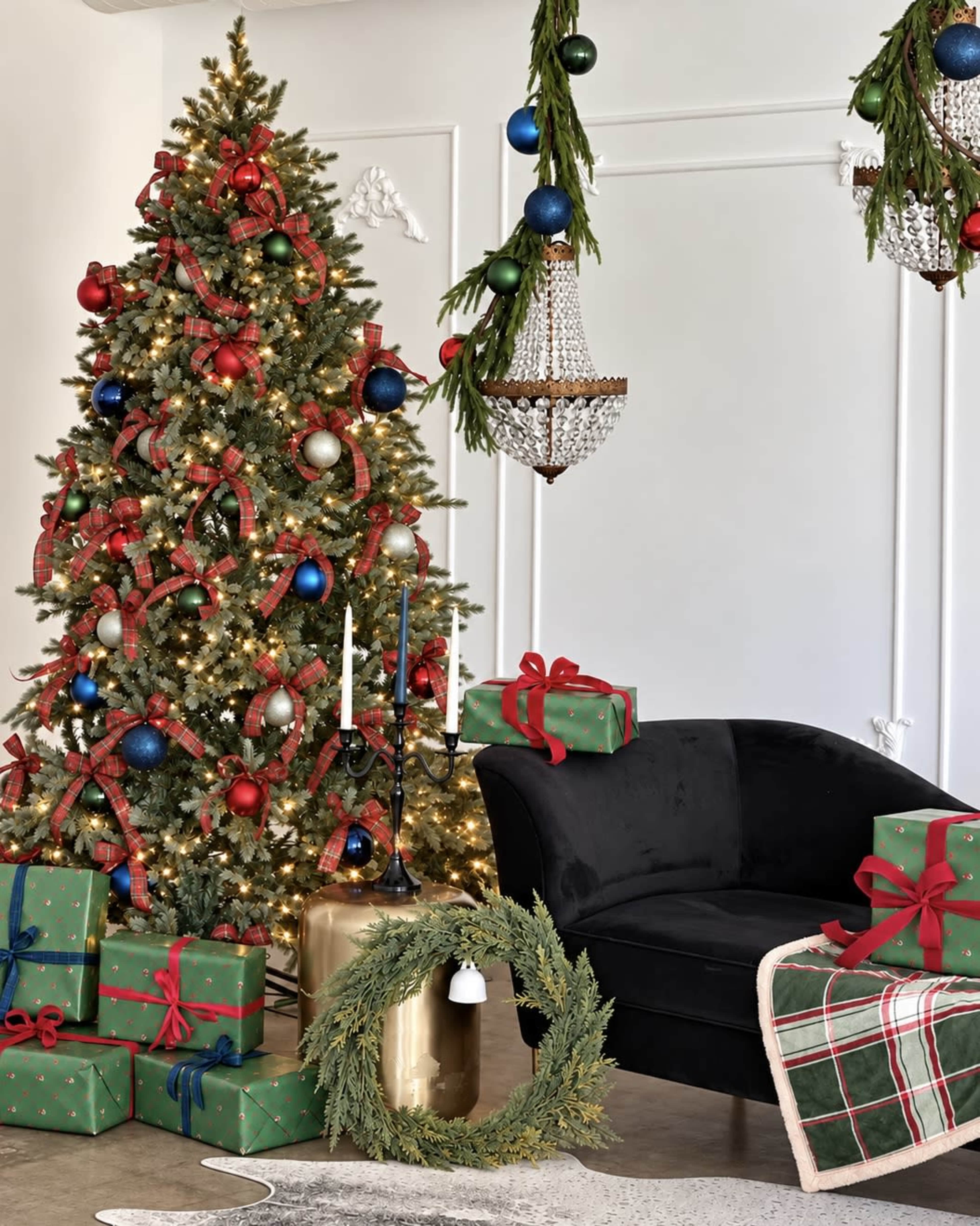 The image features a decorated Christmas tree with lights and ornaments, surrounded by wrapped gifts and a cozy black chair near a wreath and chandelier.