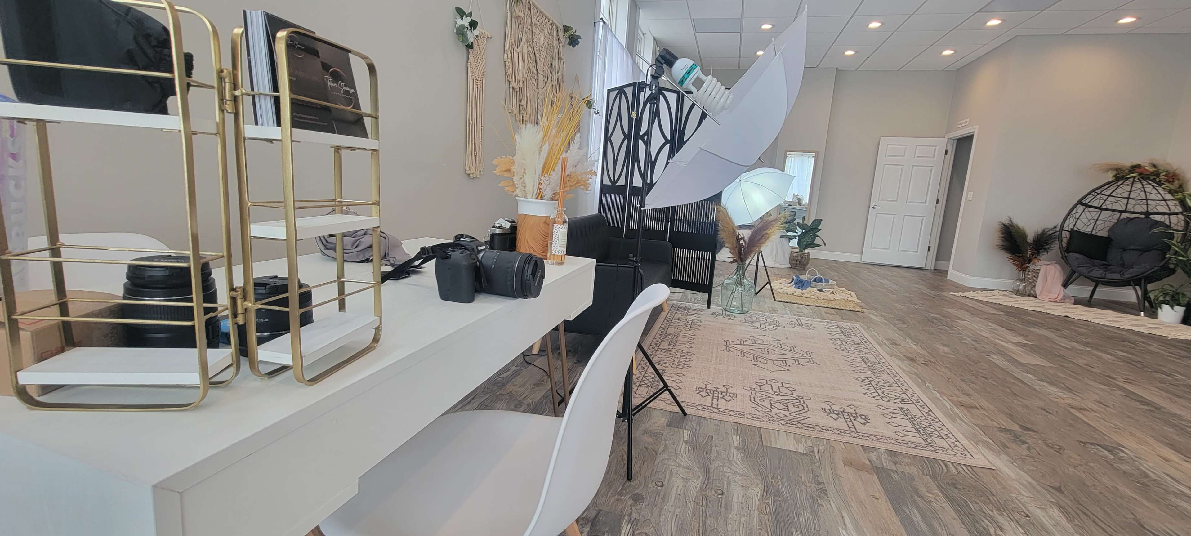 A modern studio space featuring a white desk with a camera, decorative plants, and various photo equipment, set against a light-colored wall and wooden flooring.
