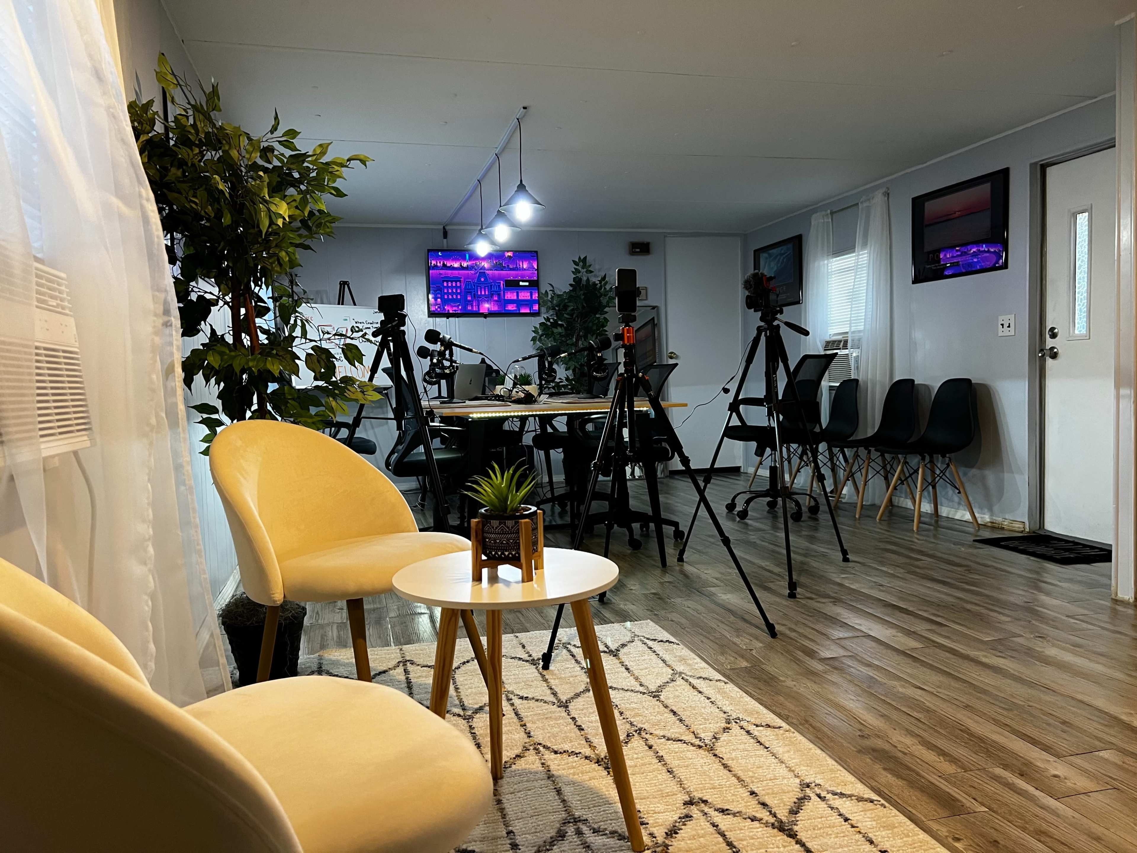 A modern room set up with a few chairs, a small table, and several cameras on tripods facing a desk with multiple monitors in a well-lit environment.