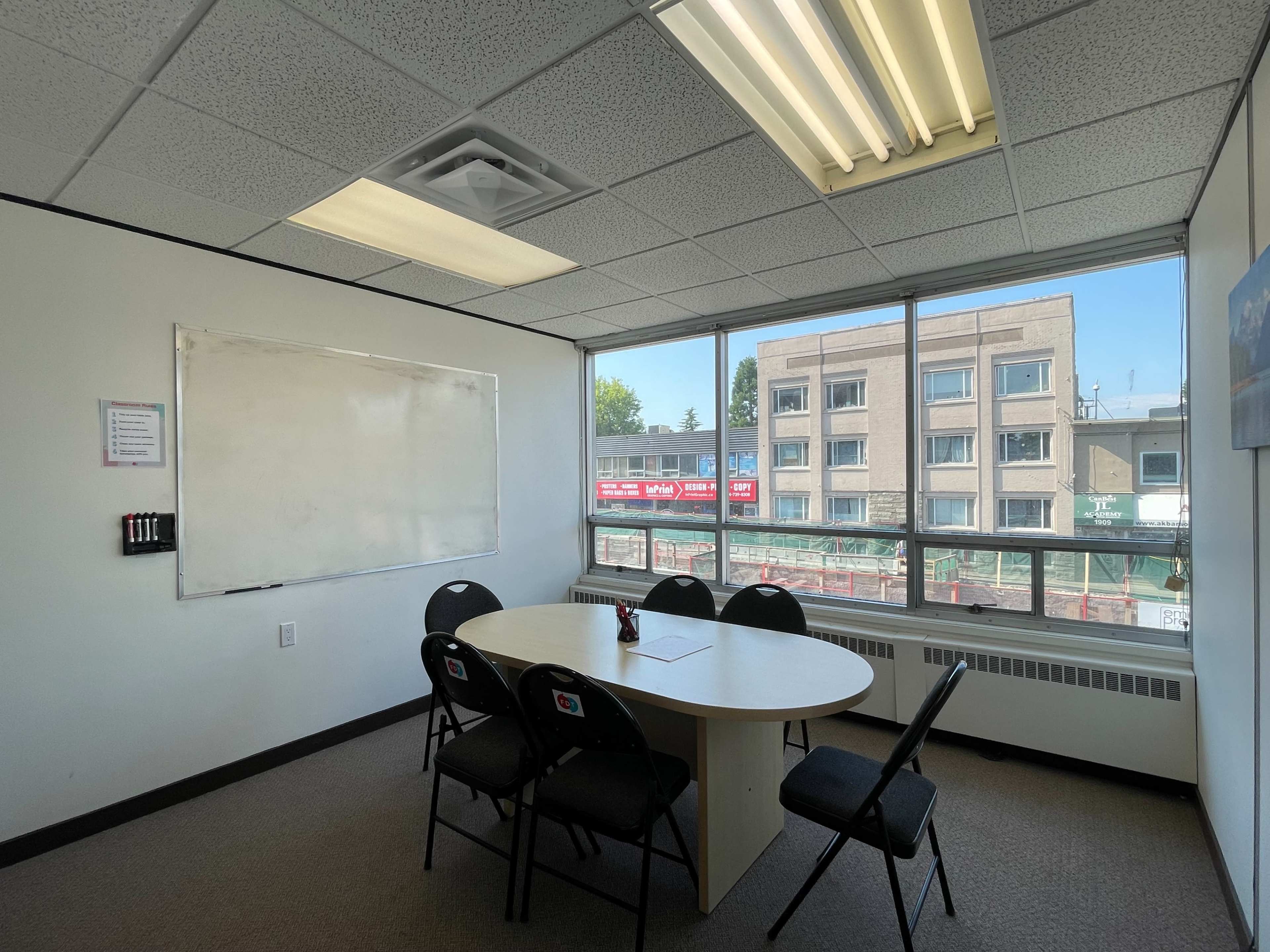A small meeting room with a round table and six chairs, large windows overlooking a construction site, and a whiteboard on one wall.