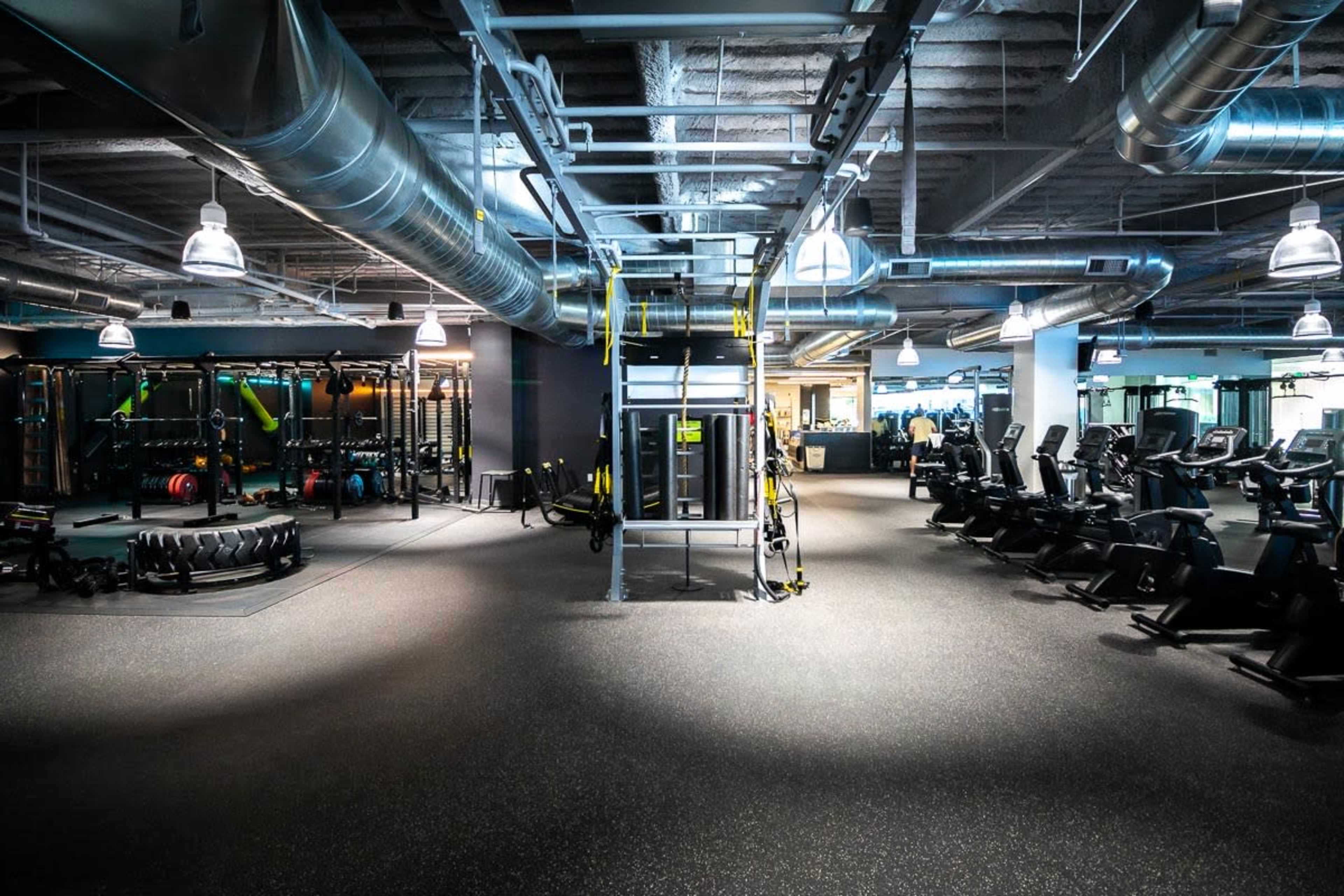 The image shows a modern gym interior equipped with various exercise machines, weights, and workout stations arranged in an open layout.