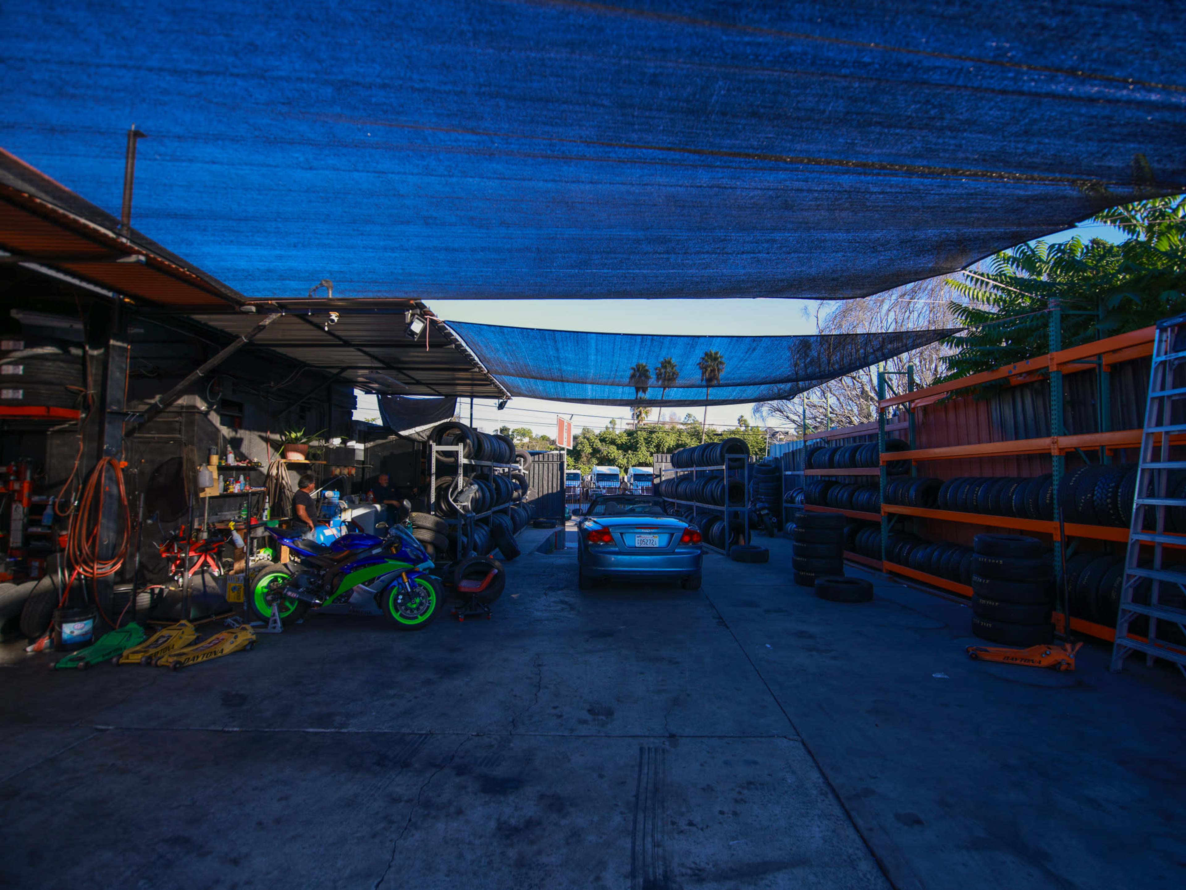 A car is parked in a tire shop with stacks of tires on both sides, covered by blue tarps overhead.