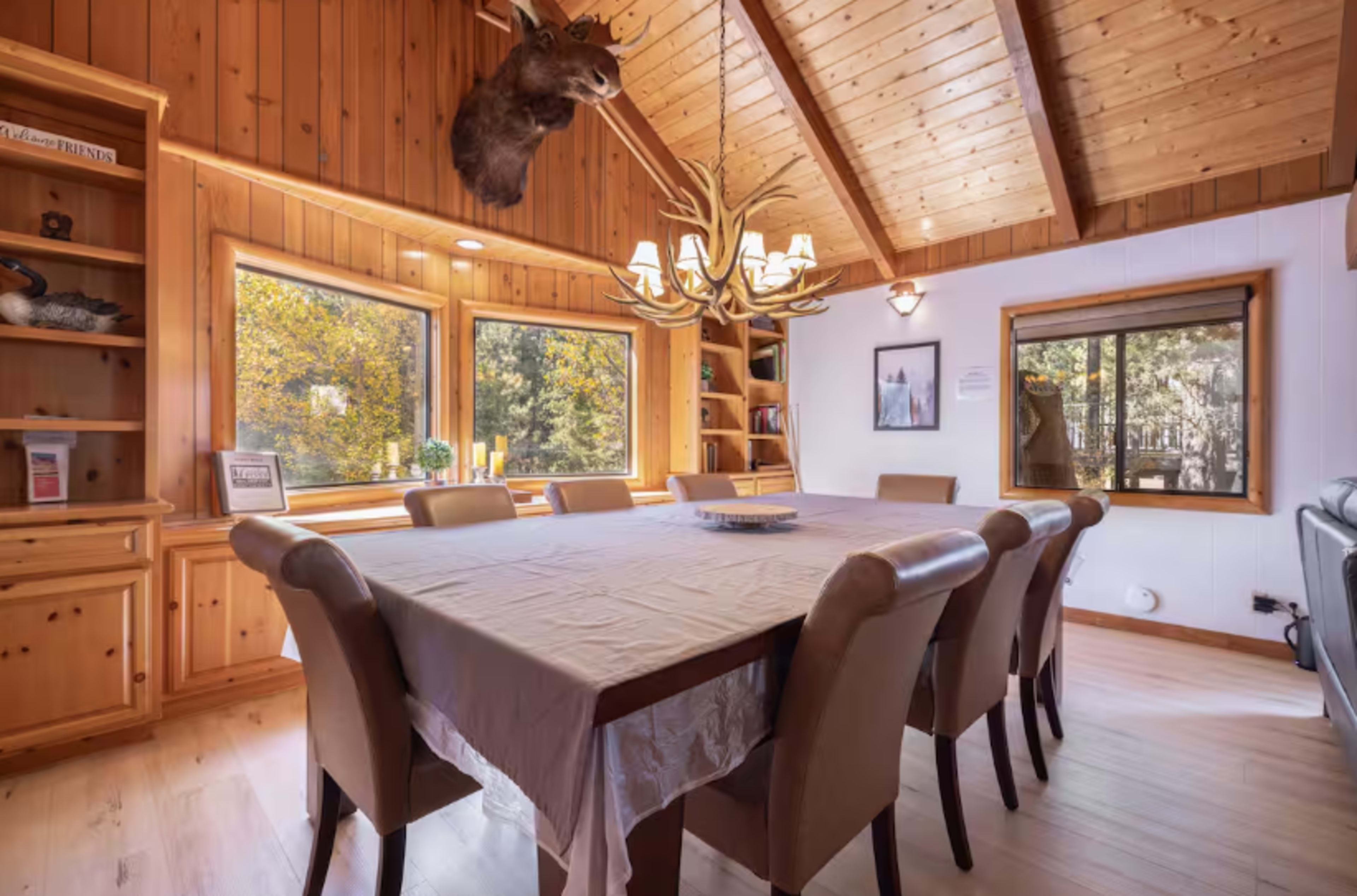 The image shows a wooden dining room with a large table surrounded by beige chairs, featuring a chandelier and large windows that let in natural light.