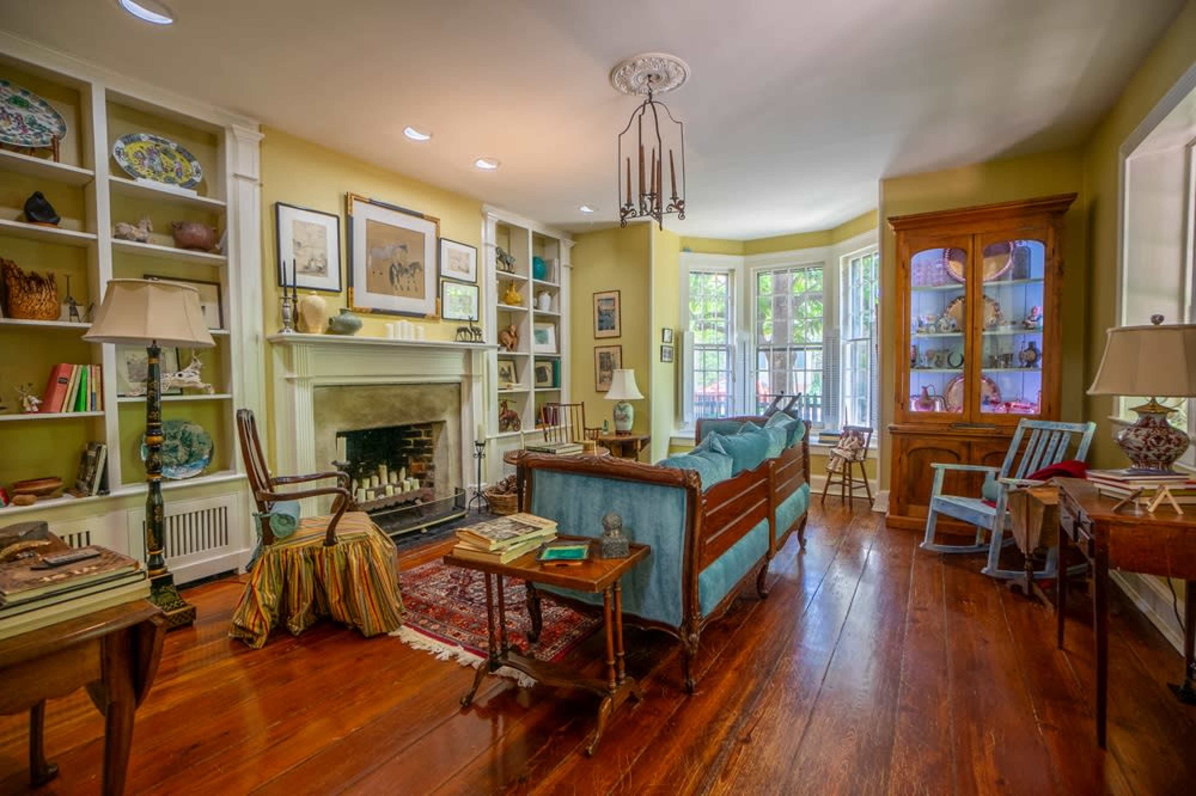 The room features a cozy living area with a fireplace, bay windows, wooden floors, and a mix of furniture and decorative items.