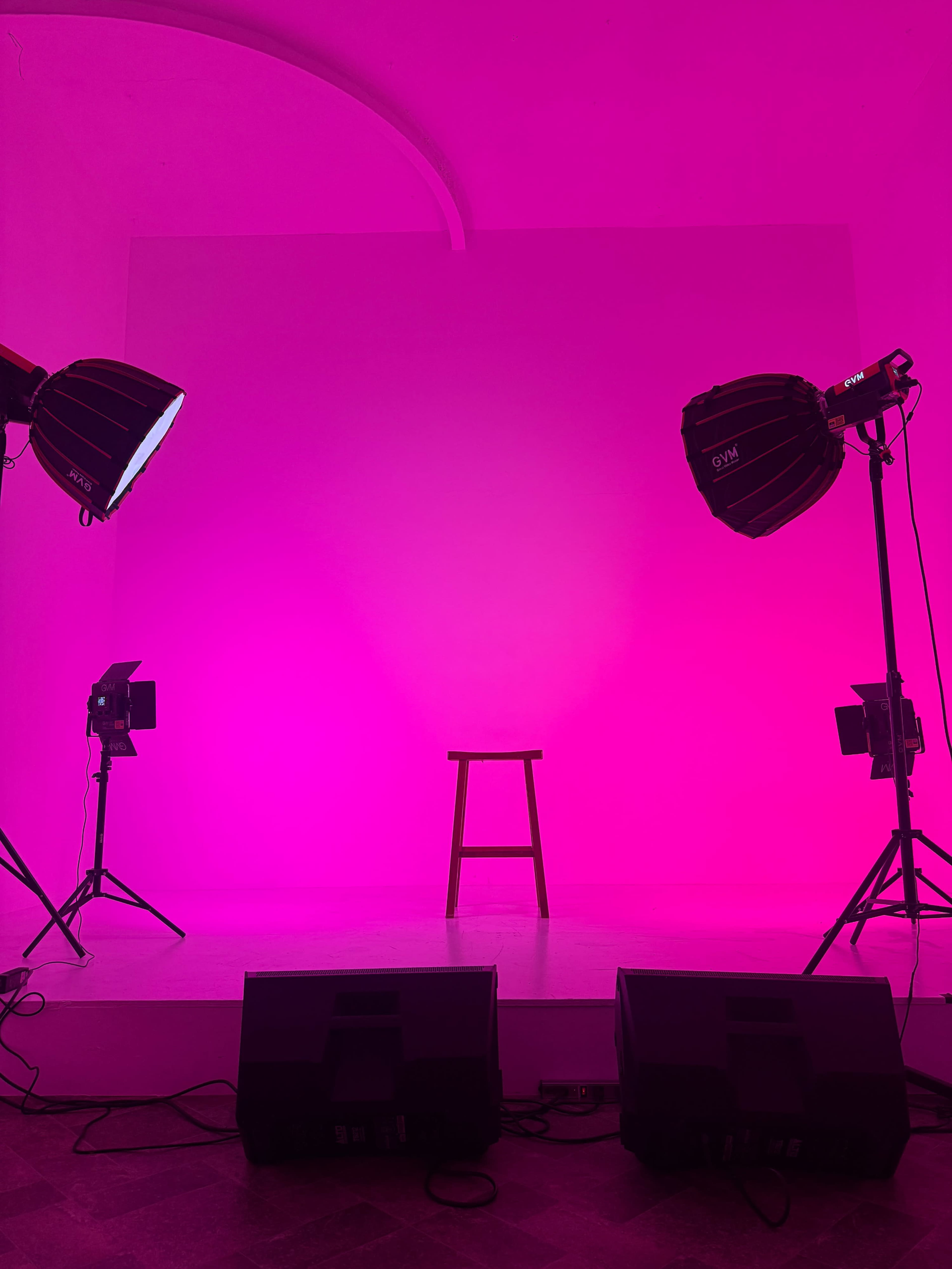 A single wooden stool is placed on a white stage illuminated by pink lighting, flanked by professional studio lights and speakers.