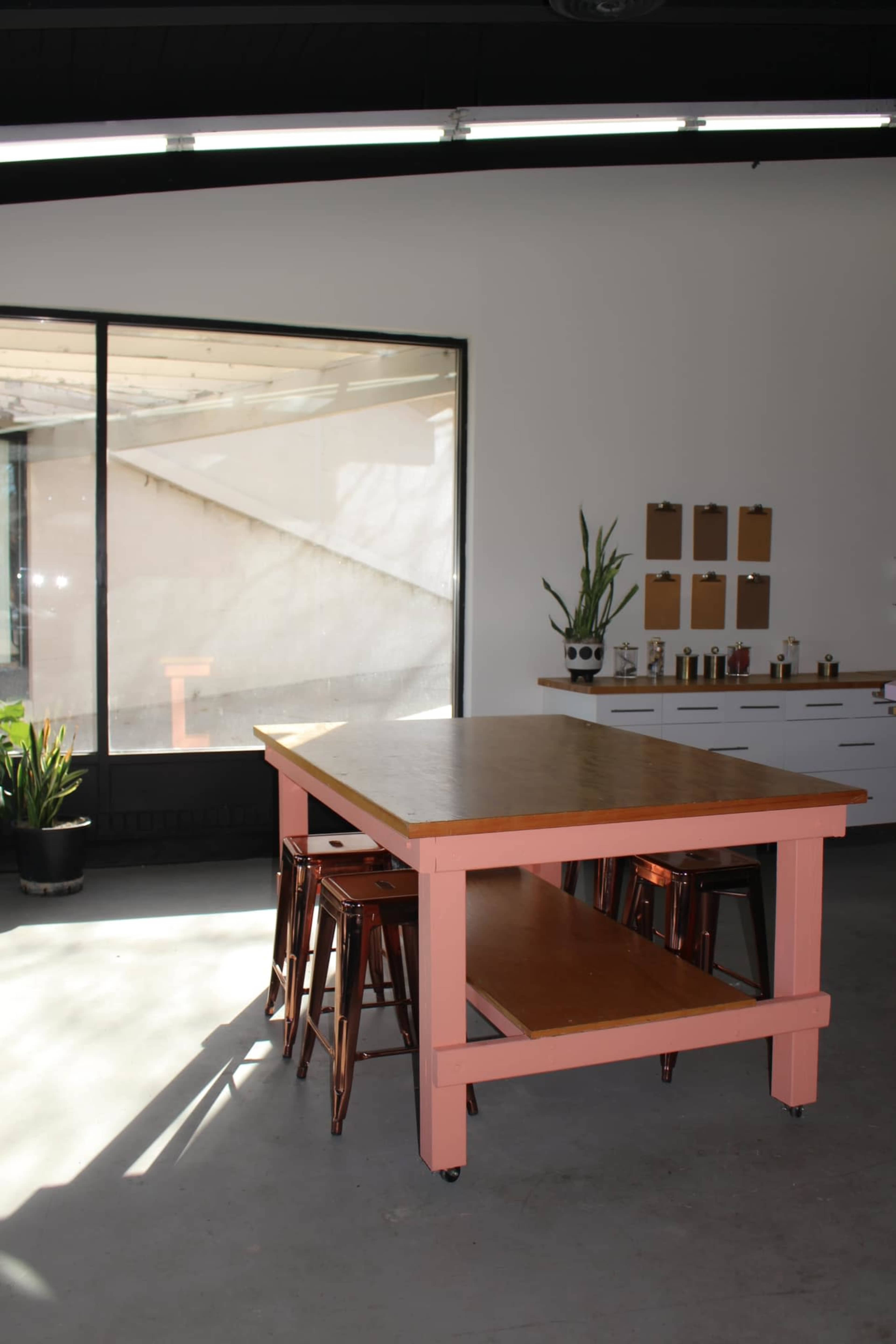 The image shows a modern kitchen space featuring a large pink table with stools, a white cabinet, and a window allowing natural light to enter.