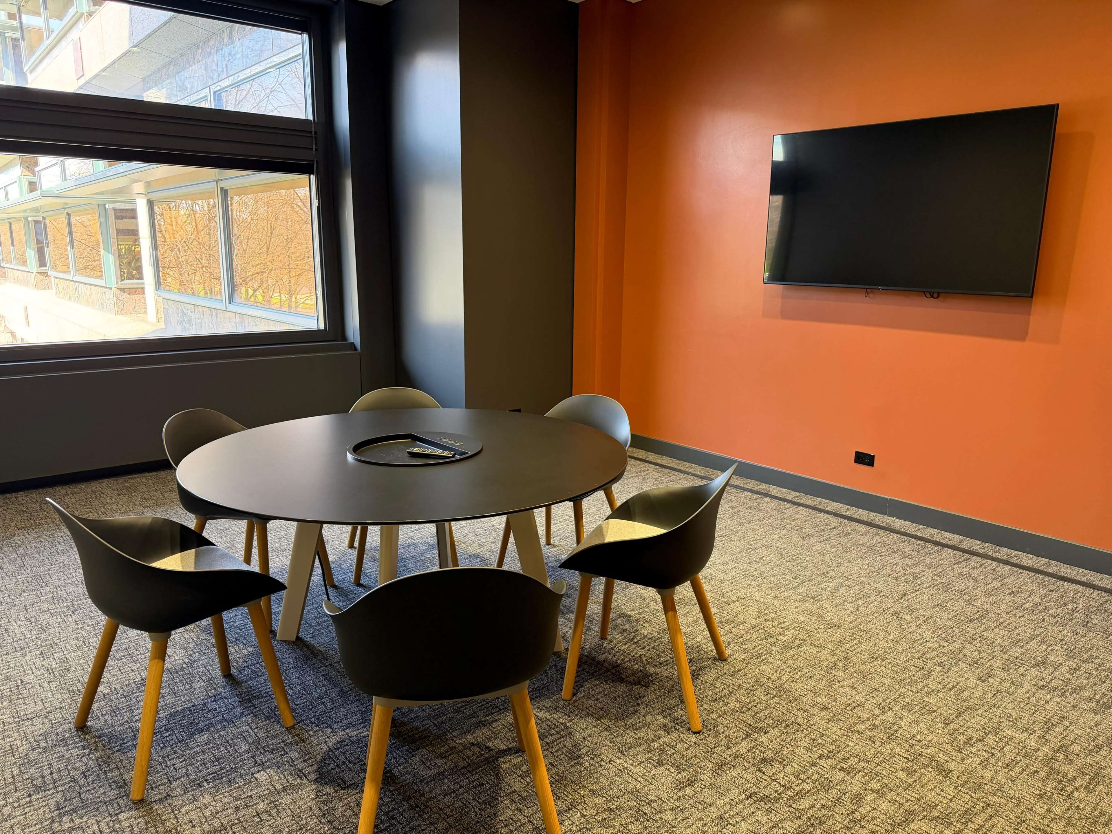 A circular table surrounded by six black chairs is positioned in a room with an orange accent wall and a large flat-screen TV mounted on the opposite wall.