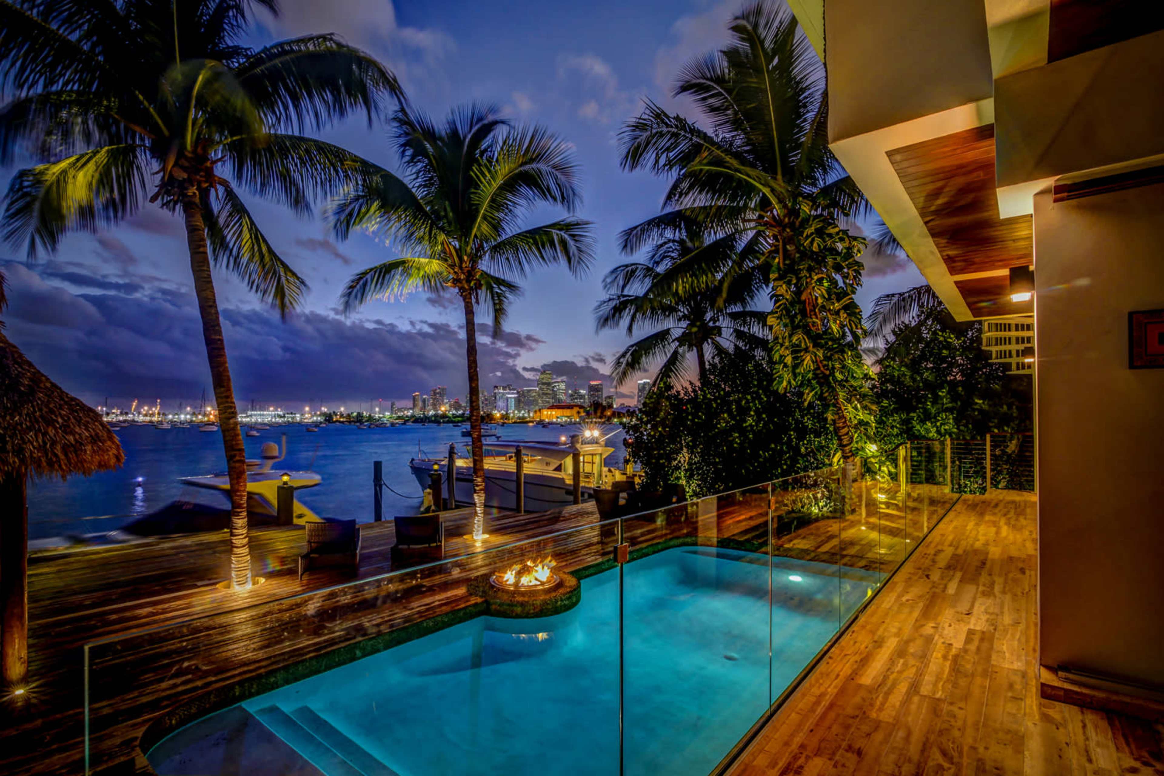 A tranquil poolside area features a glass railing, palm trees, and views of the water at dusk.