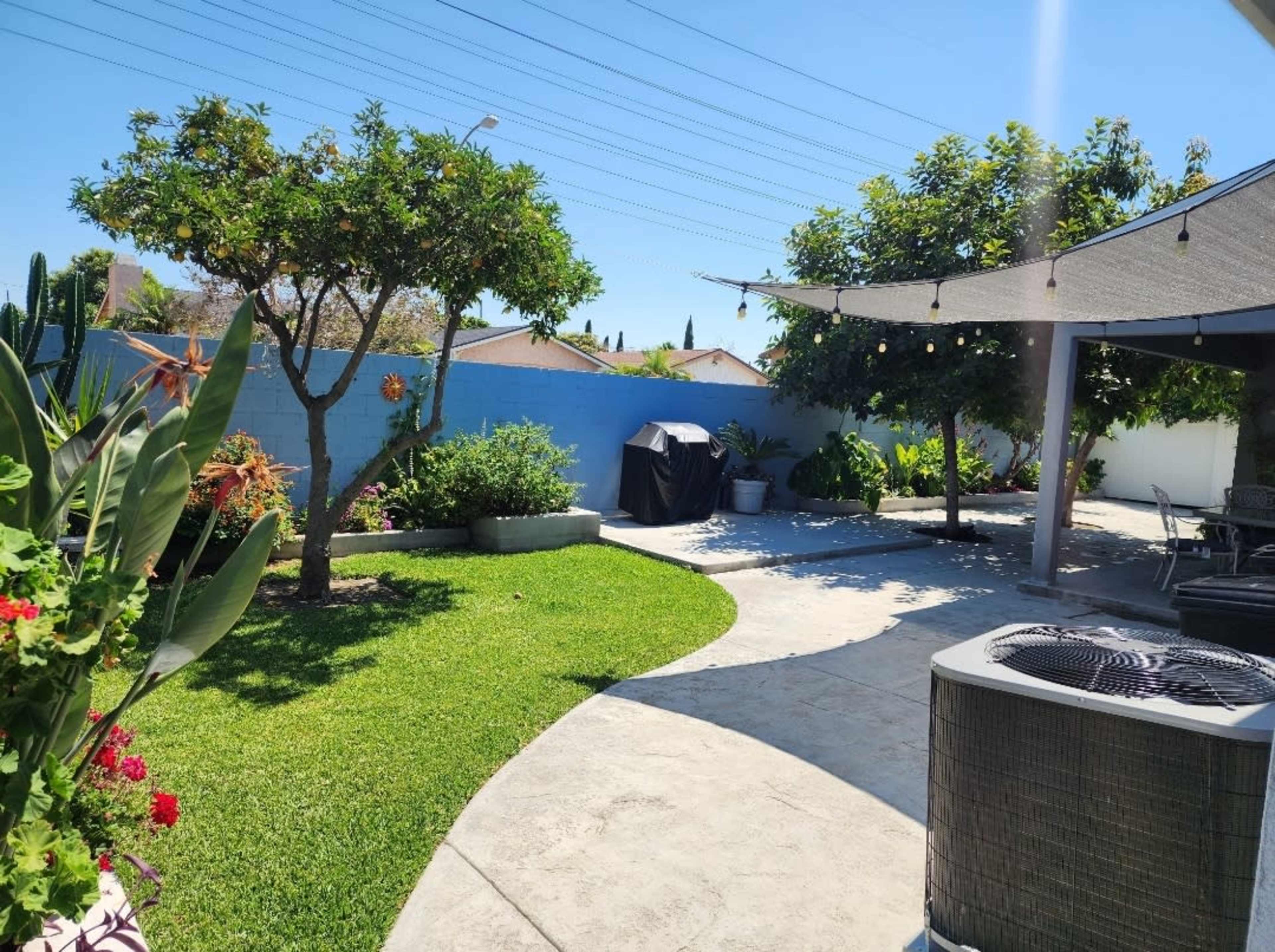 The image shows a landscaped backyard featuring green grass, trees, a barbecue grill under a cover, and a shaded seating area with outdoor furniture.