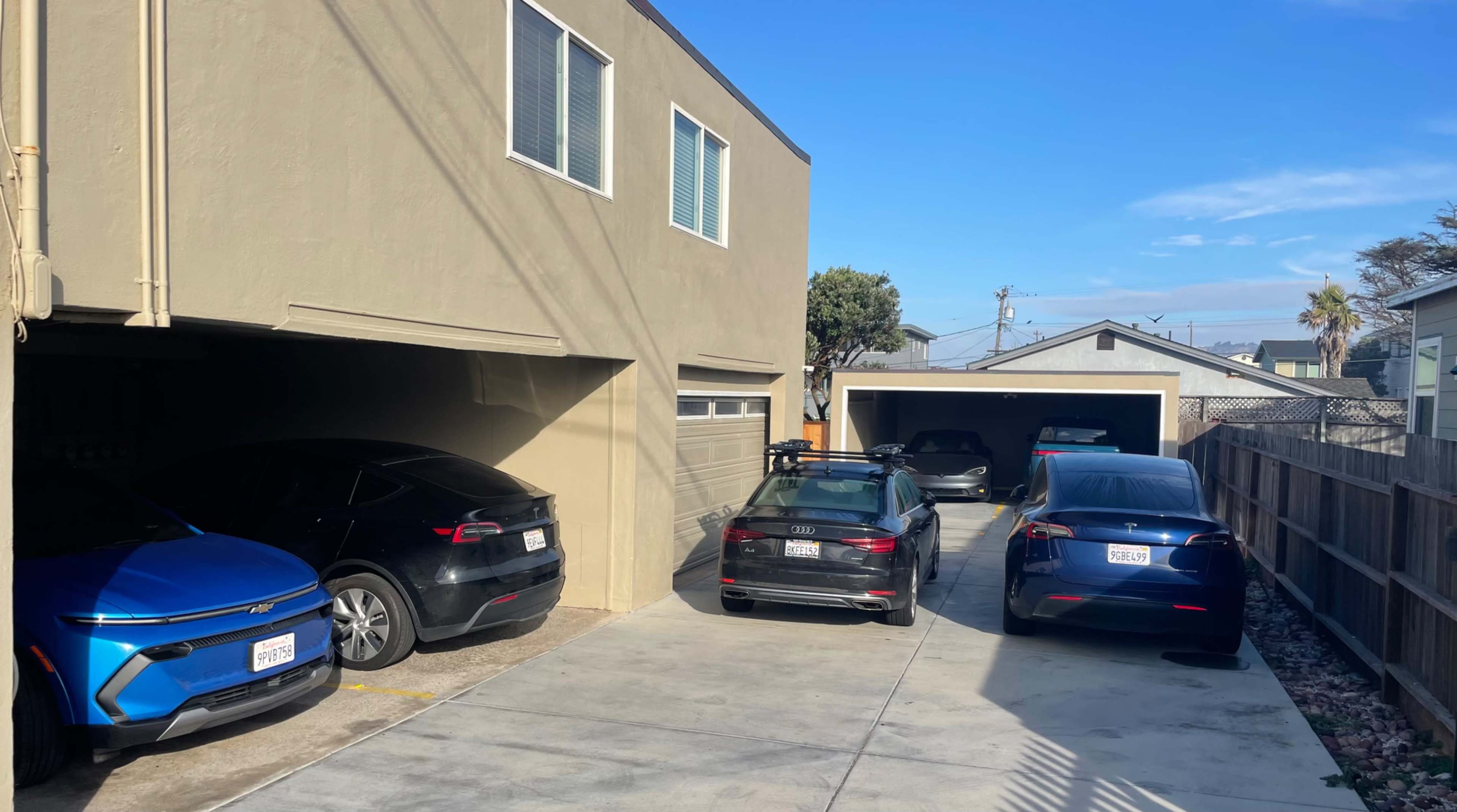 The image shows several parked cars, including two Tesla vehicles, in a residential driveway next to a beige house.