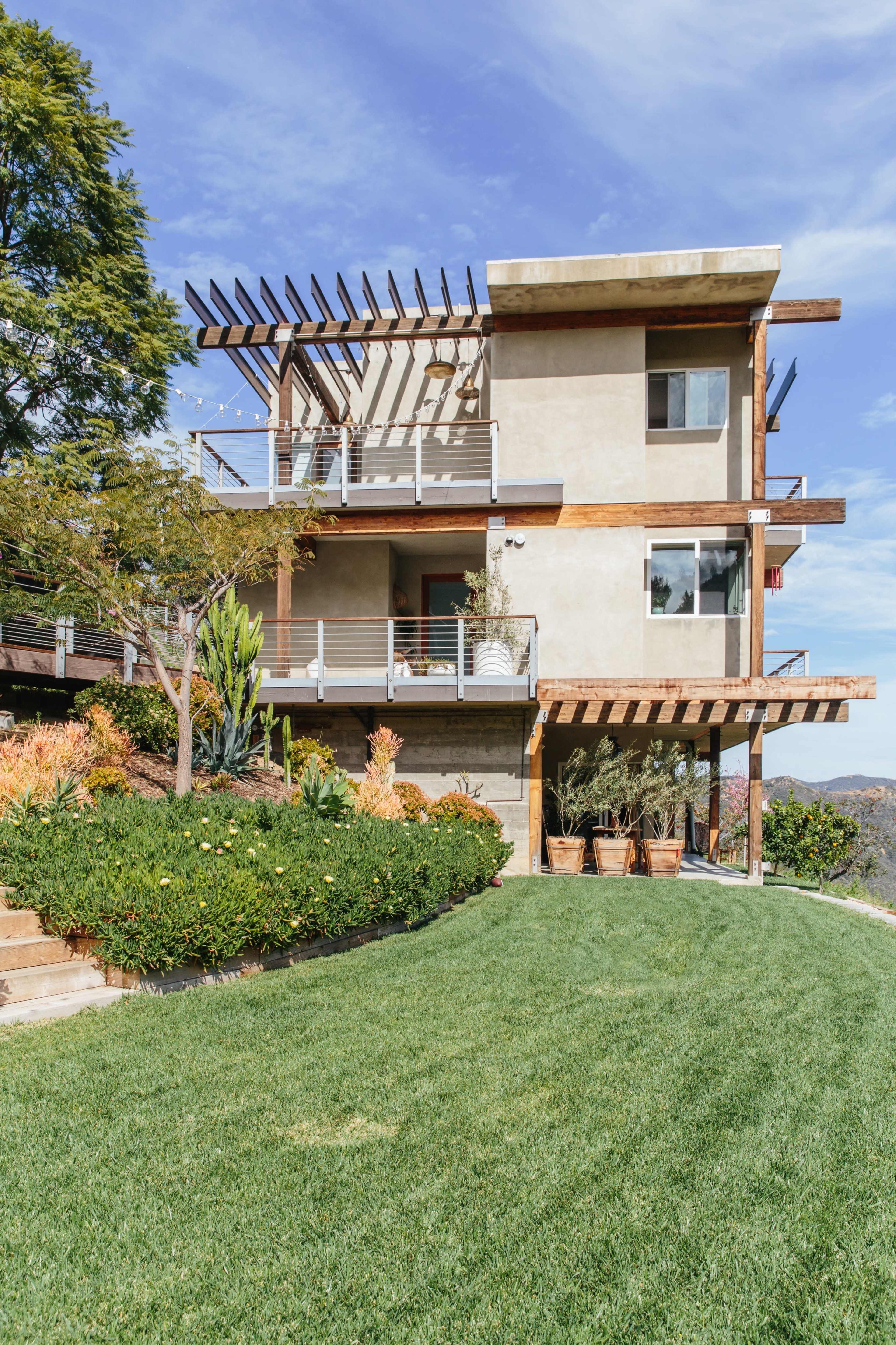 A three-story modern house with a balcony and landscaped gardens sits on a grassy hillside.