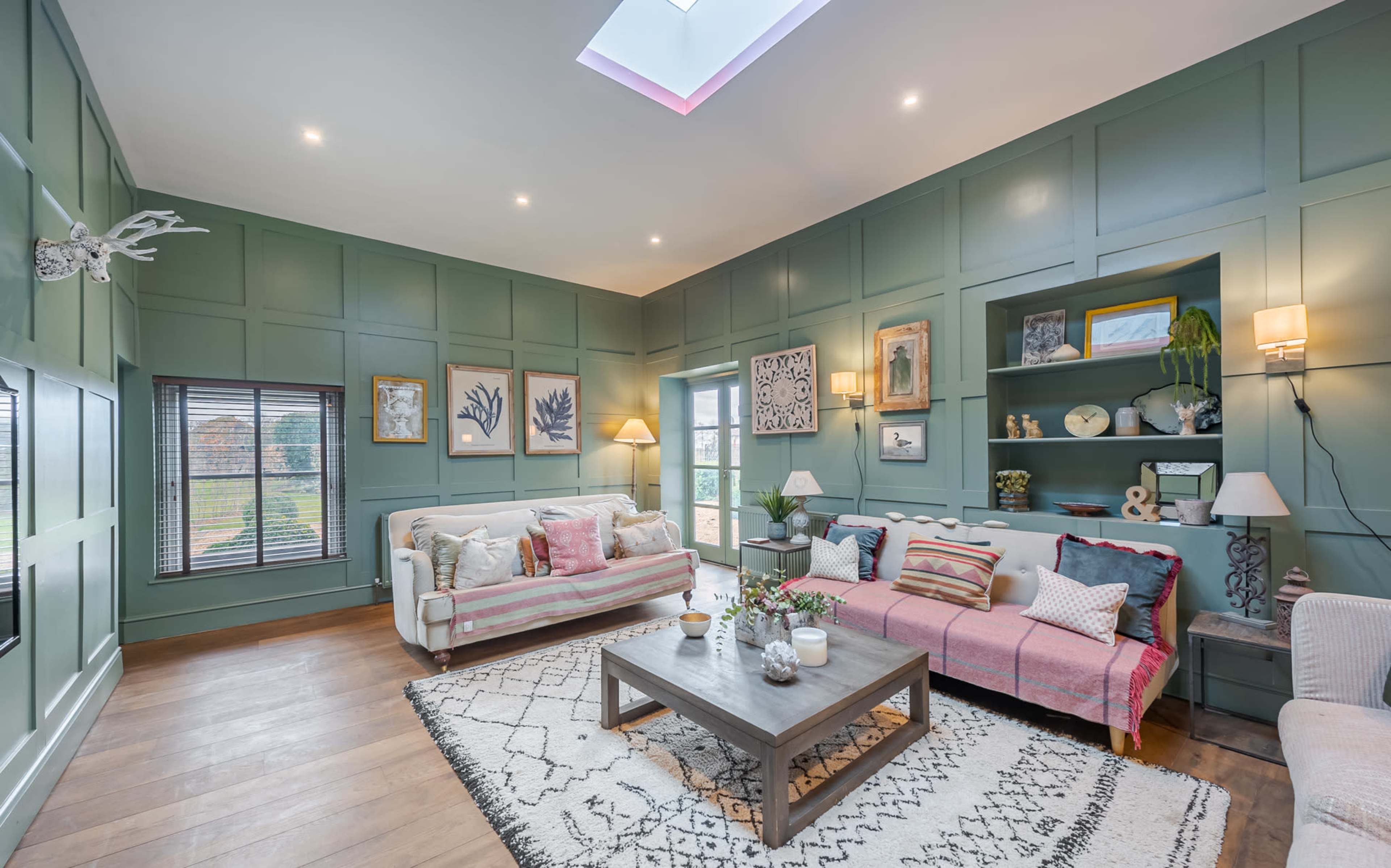 The living room features green paneled walls, a skylight, two sofas with decorative cushions, and a wooden coffee table on a patterned rug.