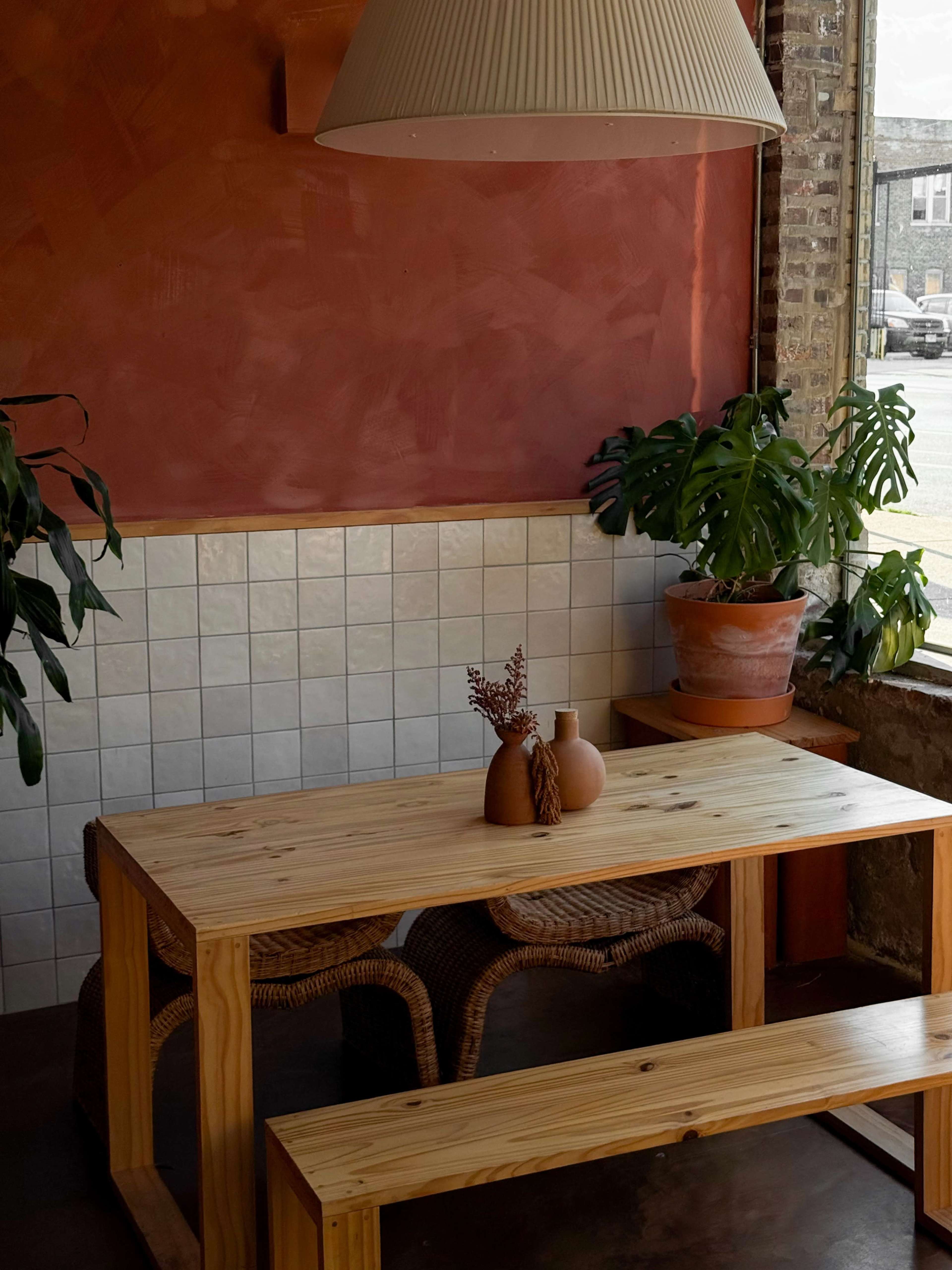 A wooden table with a bench sits in a sunlit corner, surrounded by potted plants and a textured red wall.