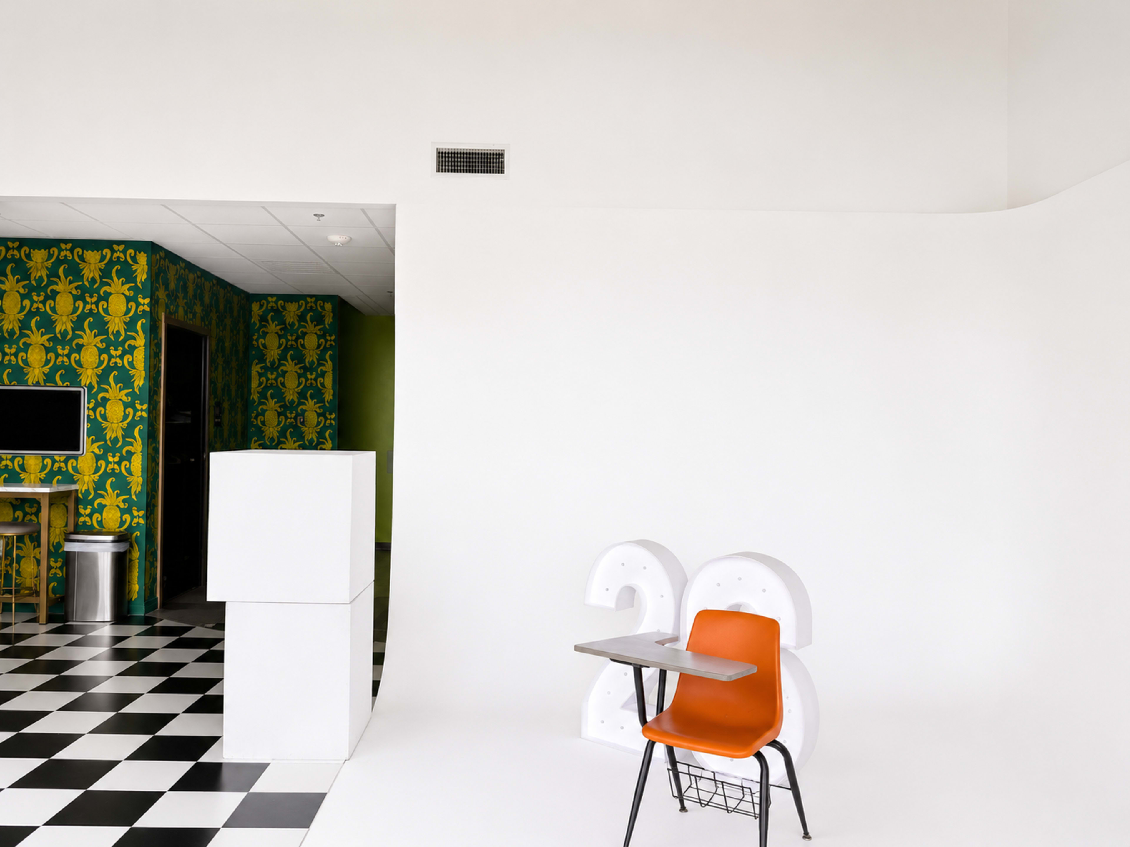 The image shows a minimalist room with a black and white checkered floor, a white wall featuring a large number "26," and an orange chair with a desk positioned in front of it.