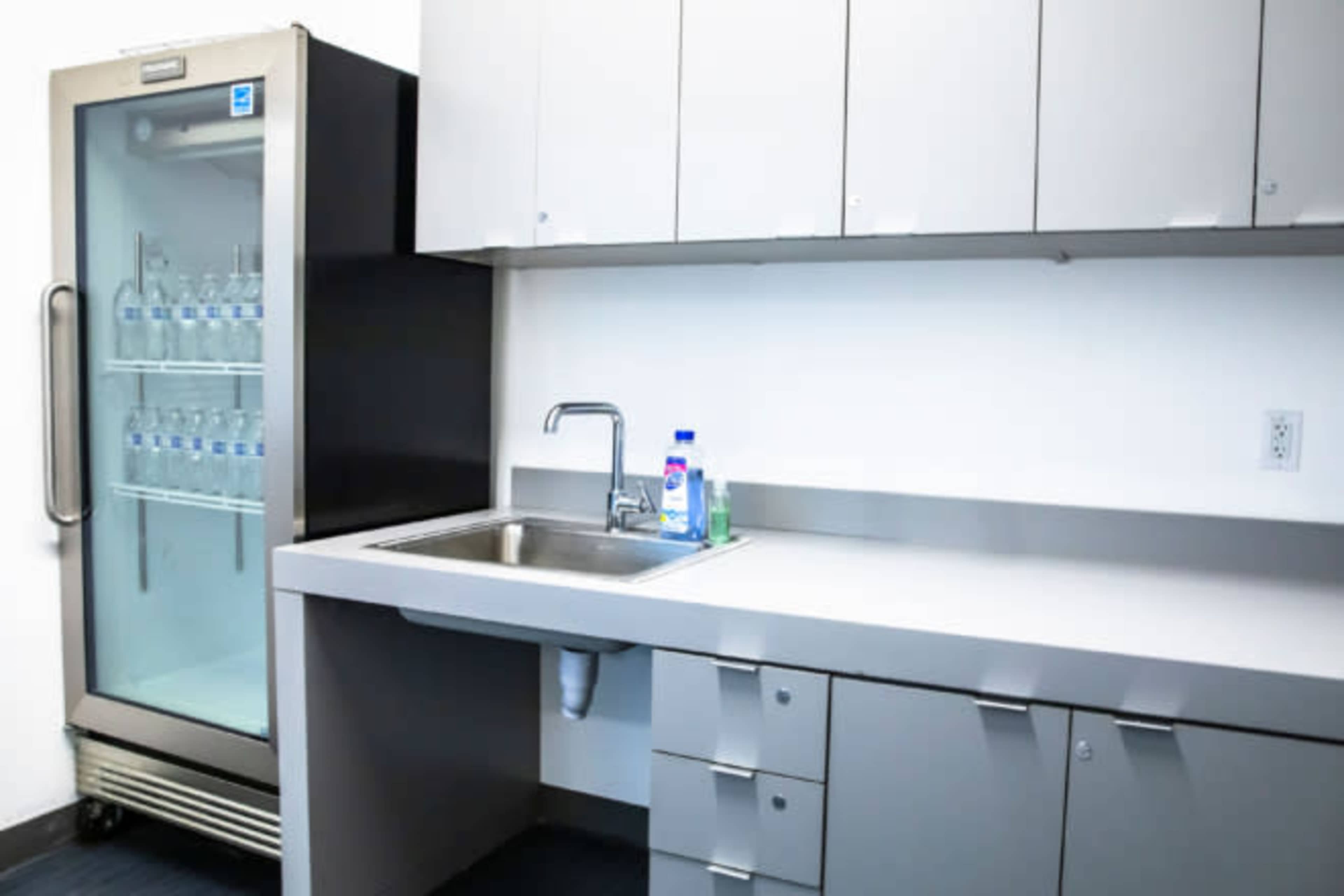 The image shows a simple kitchen space featuring a sink with cleaning supplies next to a glass-door refrigerator.