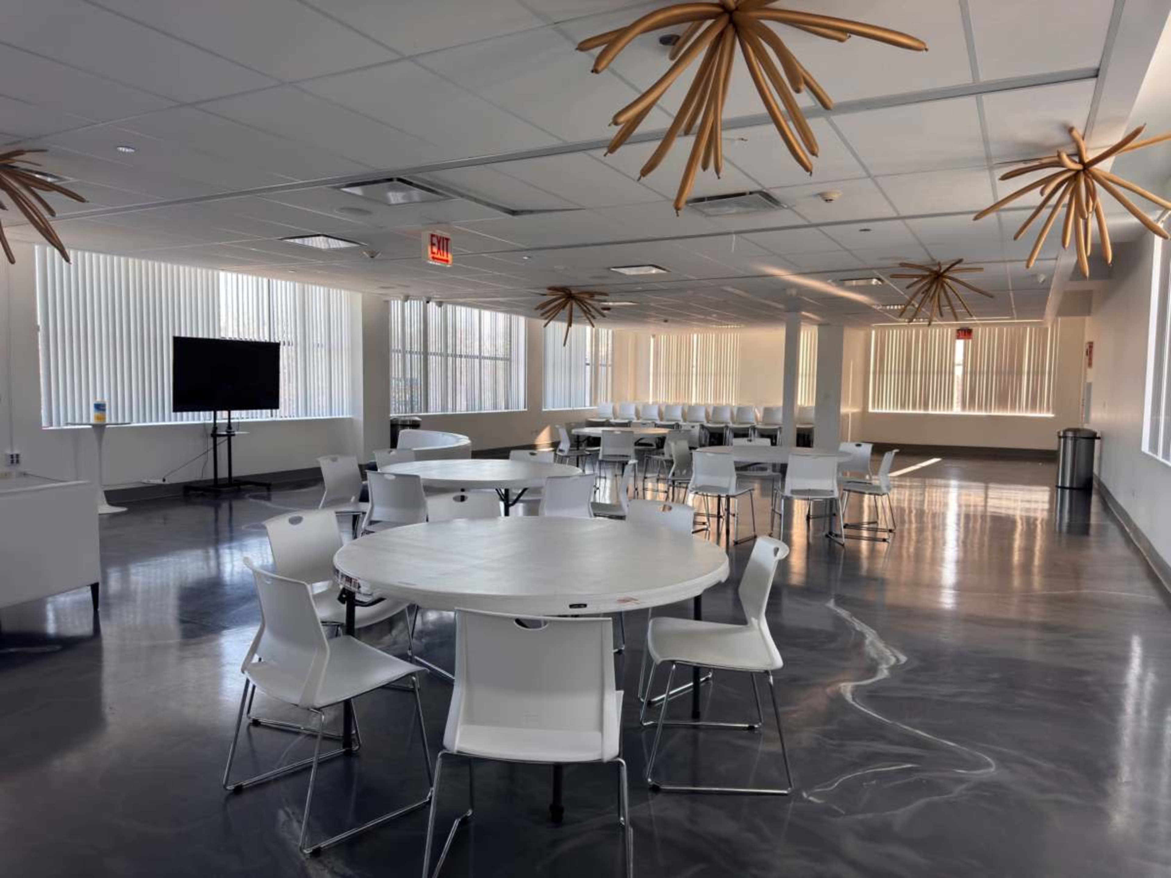 The image shows a spacious, modern office break room with round tables and white chairs, illuminated by natural light from large windows and featuring unique ceiling decorations.